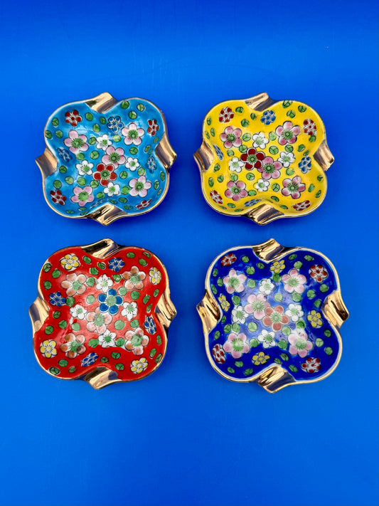 Four square ceramic ashtrays with vibrant floral patterns in teal, yellow, red, and navy blue, arranged in a grid, blue background