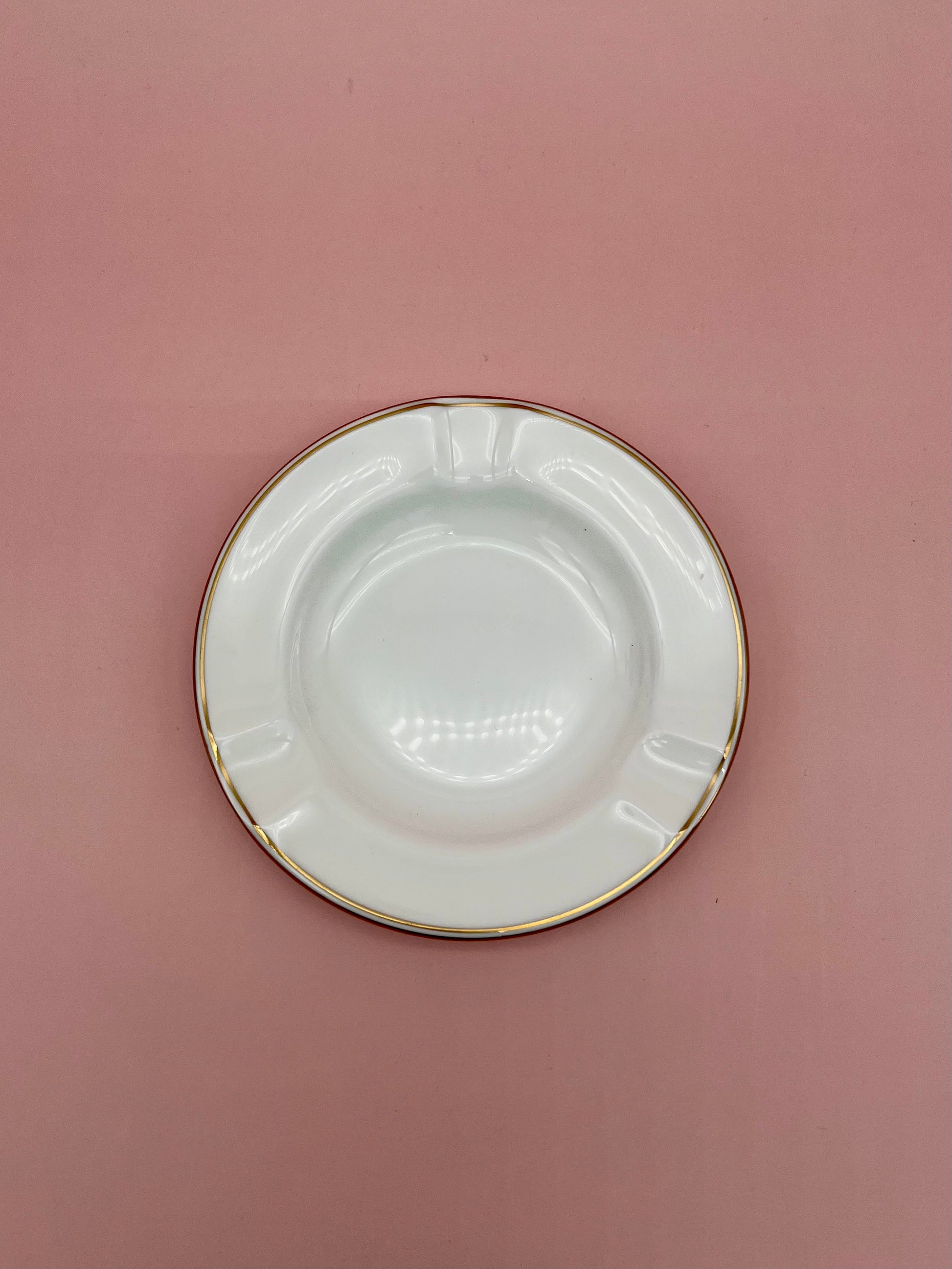 Round white ceramic ashtray with a thin gold rim, dusty rose background.