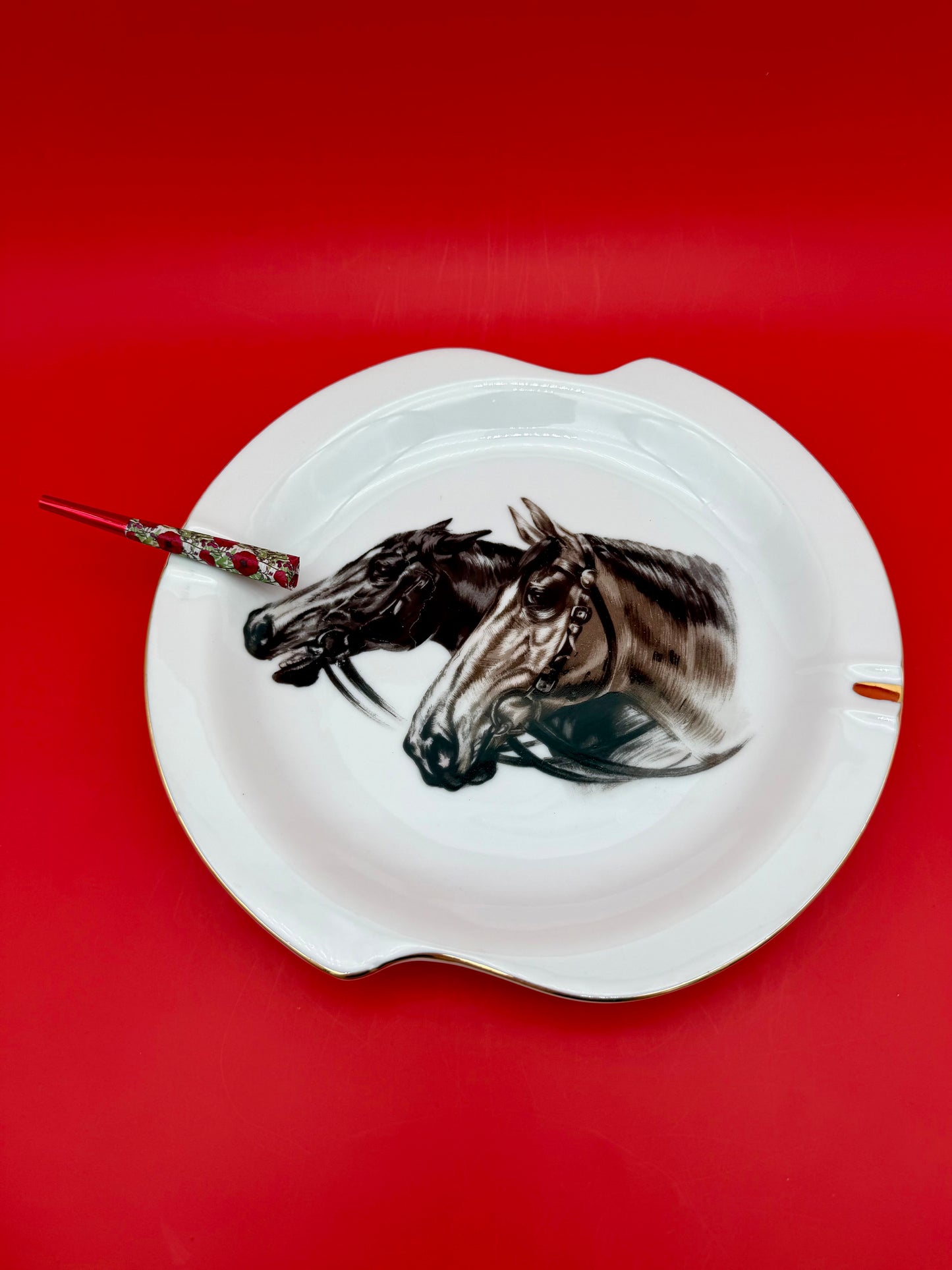 White scalloped ashtray from Duke with a close-up horse head portrait, glass cigarette pick resting across it, red background.