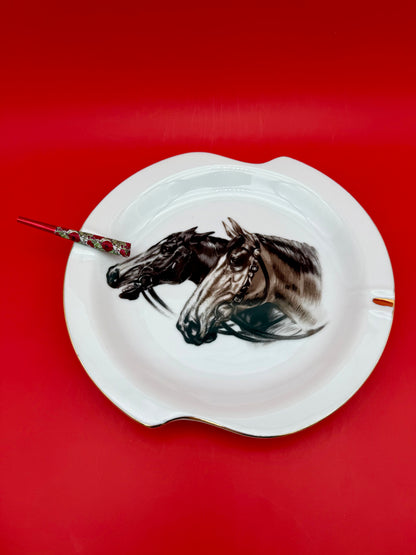 White scalloped ashtray from Duke with a close-up horse head portrait, glass cigarette pick resting across it, red background.