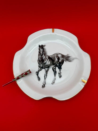 White scalloped ceramic ashtray hand-painted with a black and white horse, glass cigarette pick resting across it, red background.