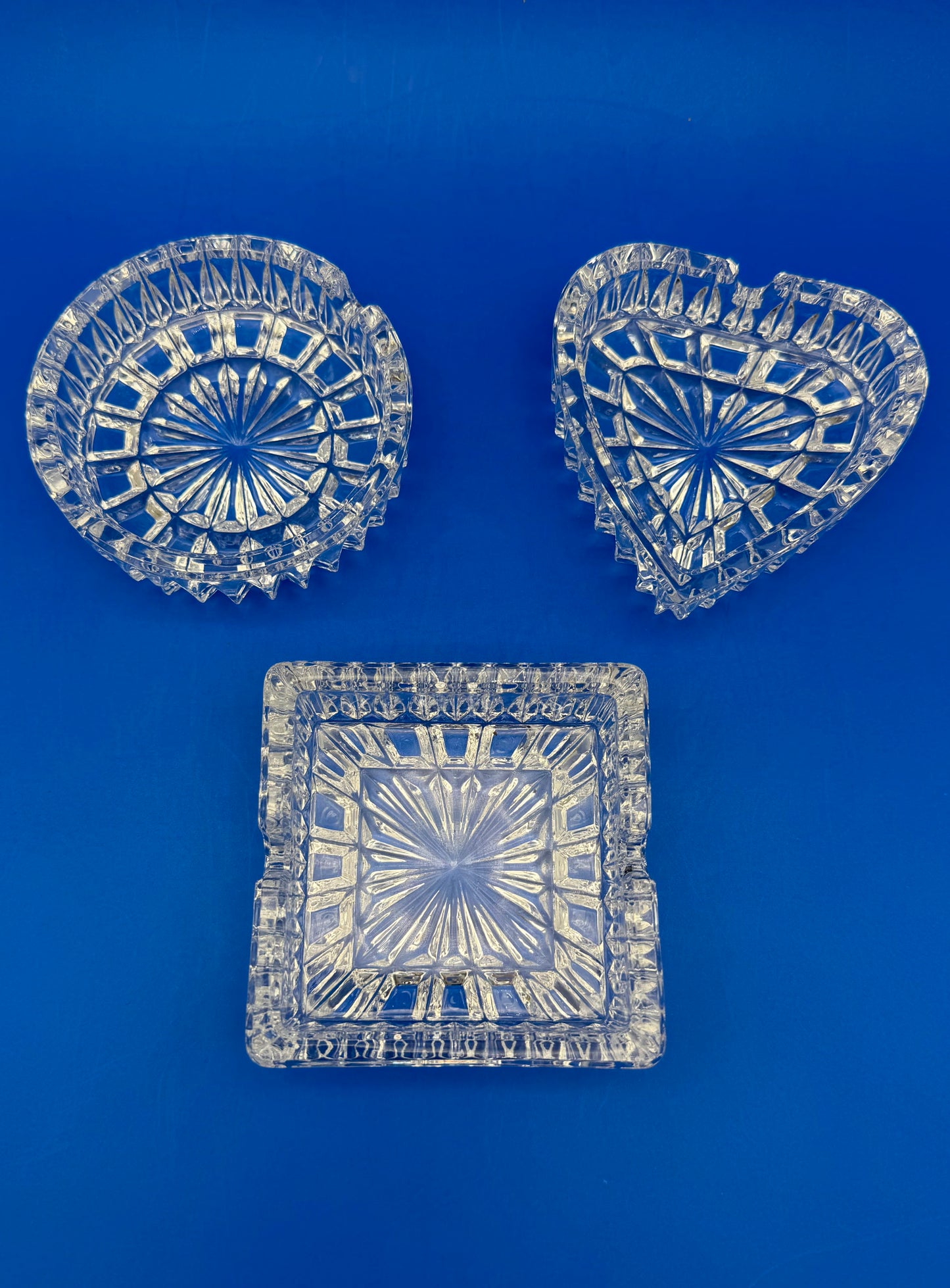Round, heart-shaped, and square faceted crystal ashtrays with starburst patterns arranged together, blue background