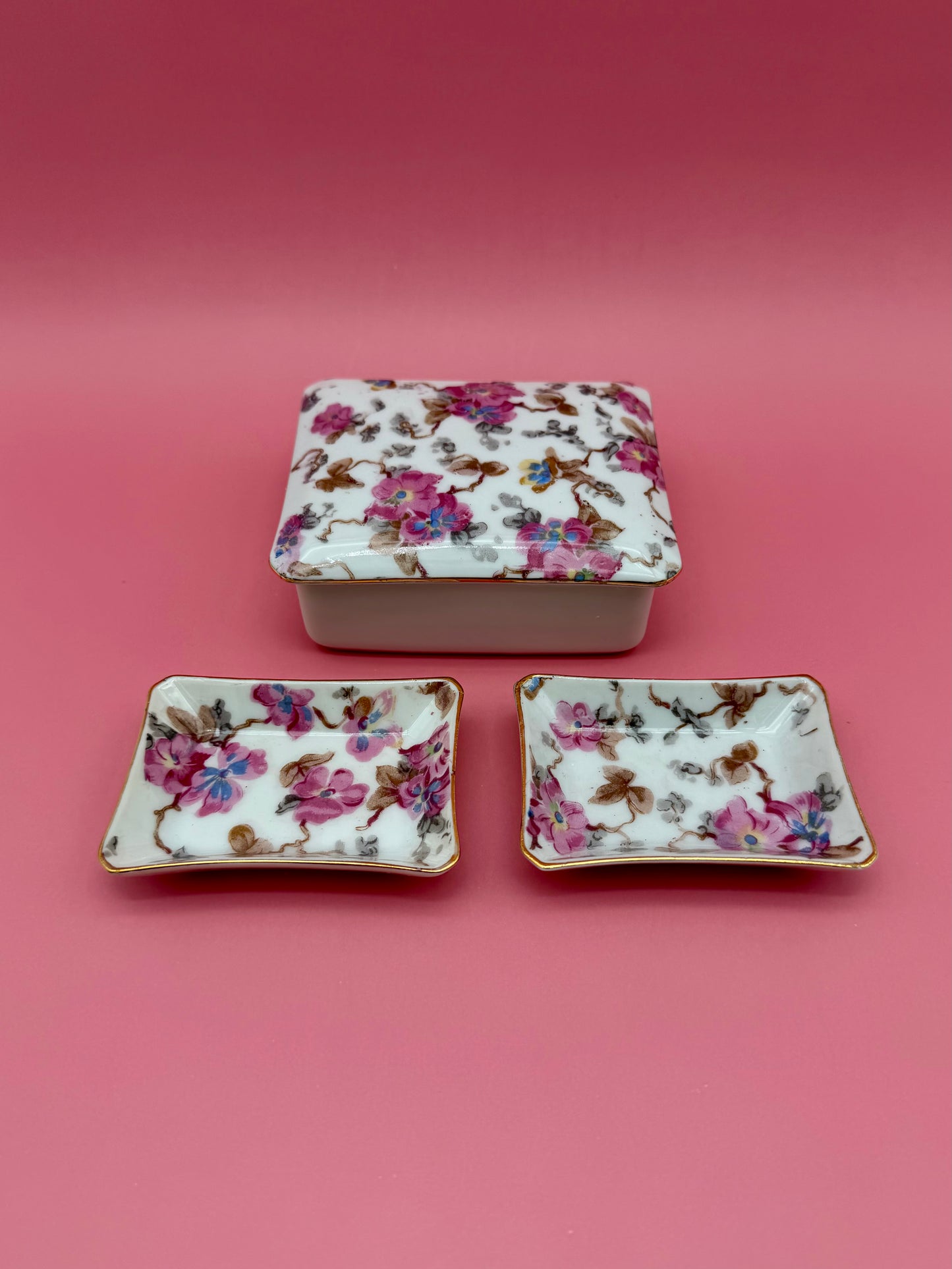 Closed floral porcelain stash box stacked above two matching floral ashtrays, dark pink background