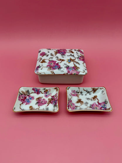Closed floral porcelain stash box stacked above two matching floral ashtrays, dark pink background