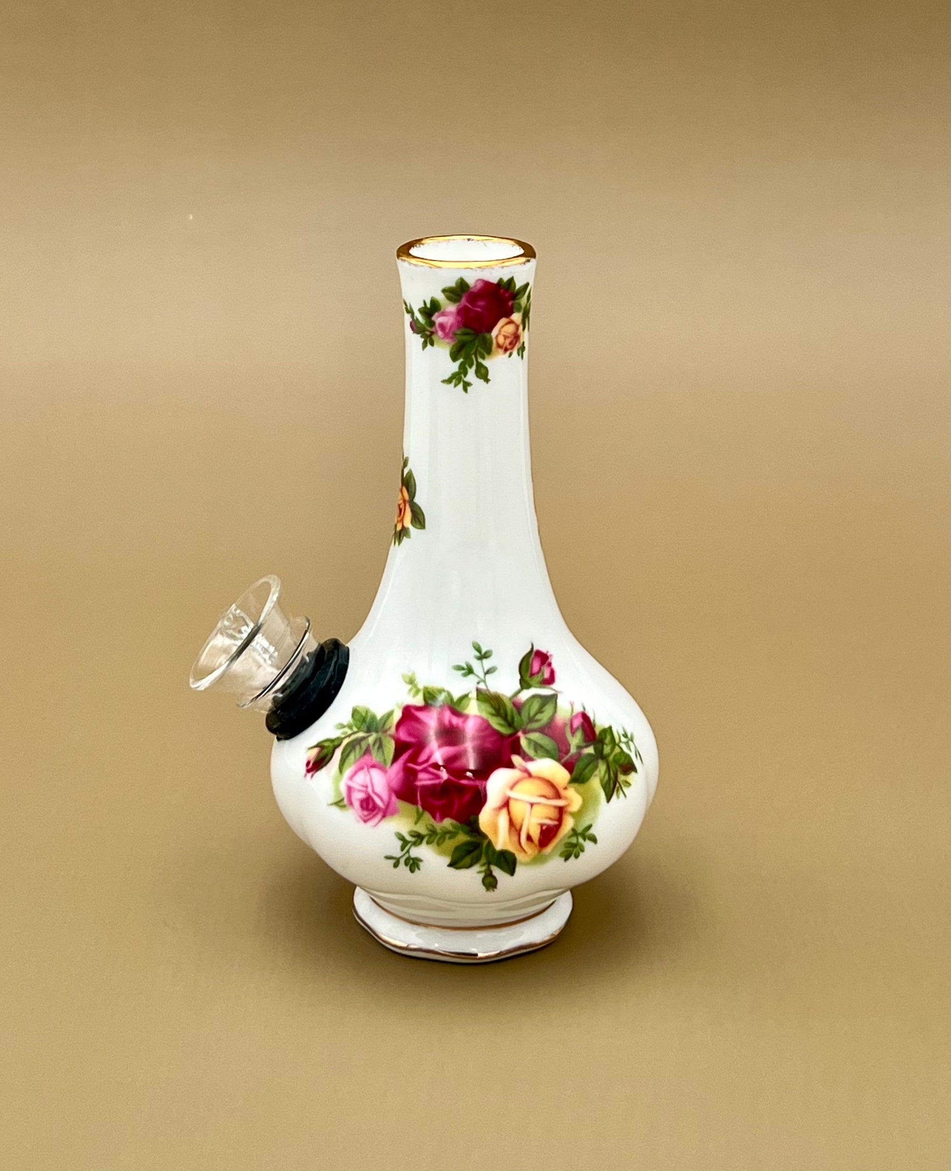 Front view of a white porcelain bud vase water pipe with a hand-painted bouquet of red and pink roses and gold trim, glass bowl piece on the side, warm gold-tan background.