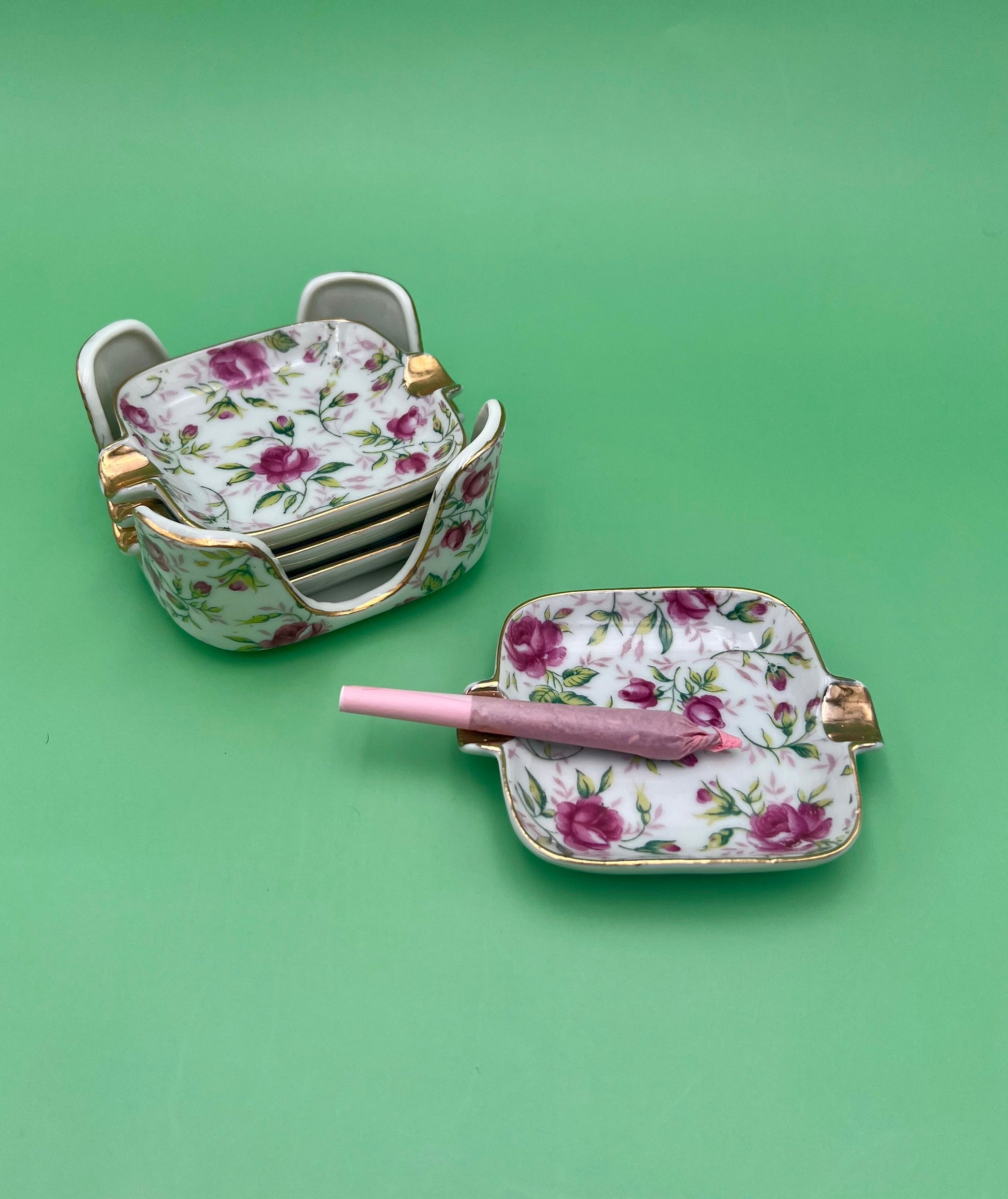 Stack of square rose-patterned ashtrays with gold handles from the Elaine Set, beside a single matching ashtray with a glass cigarette pick with a pink handle resting across it, green background.