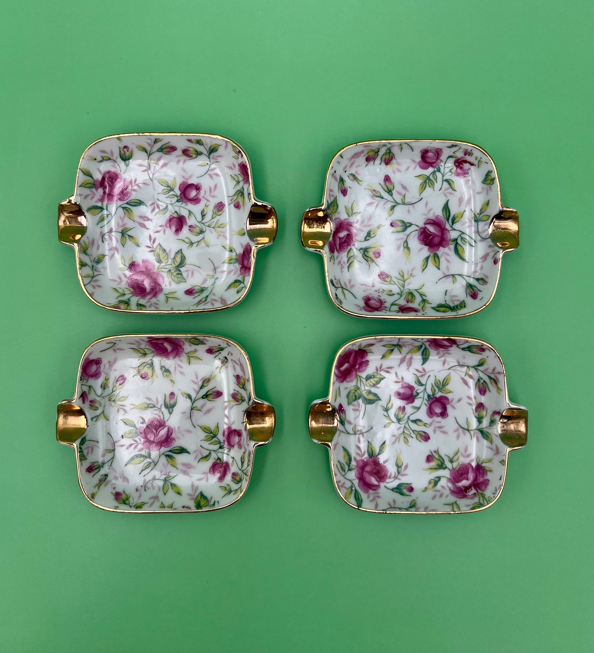 Four square rose-patterned ashtrays with gold handles from the Elaine Set arranged in a 2x2 grid, green background.