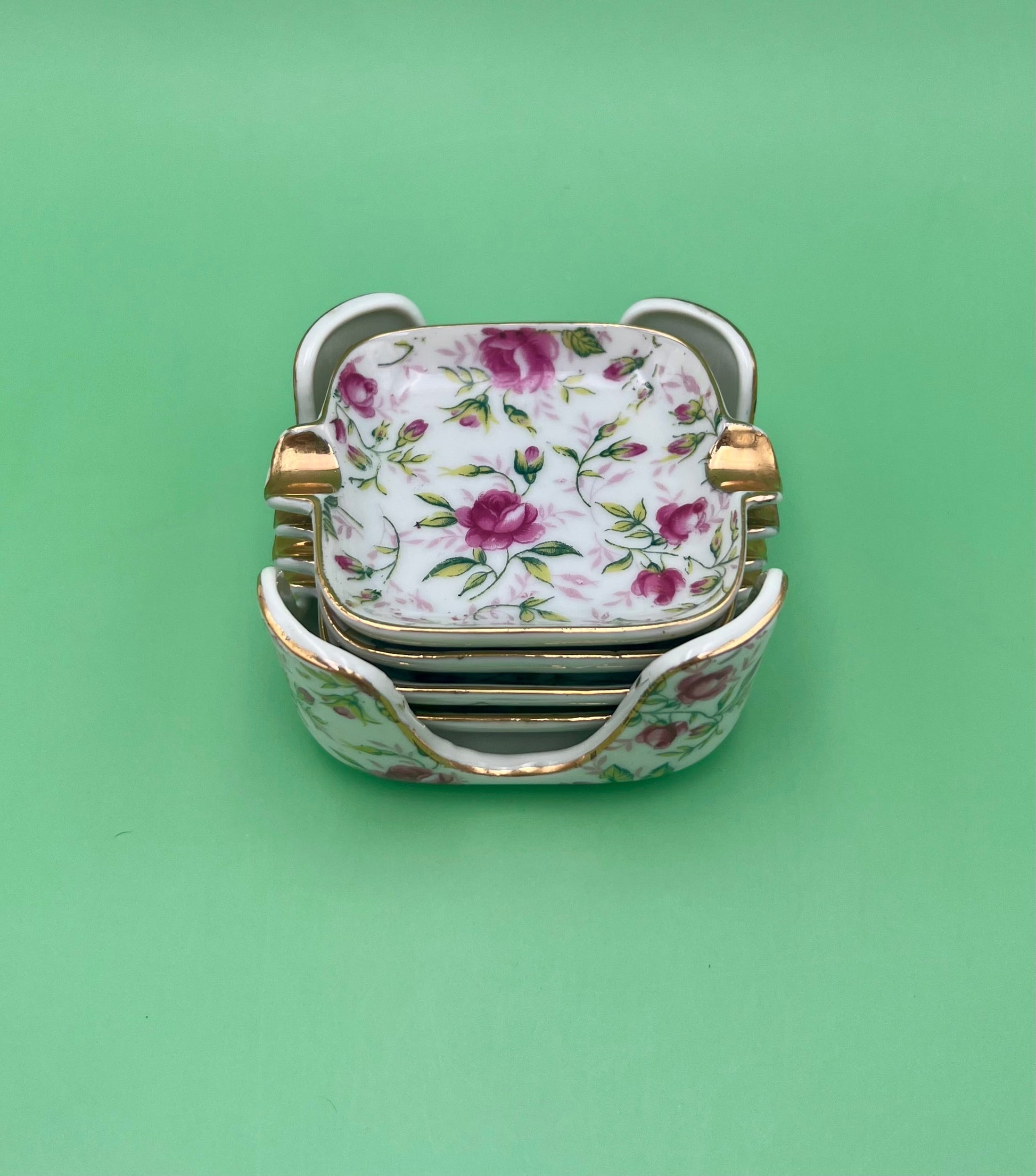 Stack of square porcelain ashtrays from the Elaine Set with a vintage rose pattern and gold trim, green background.