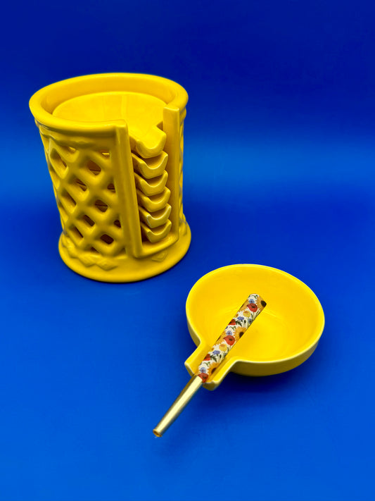 Yellow ceramic lattice ashtray holder beside a round yellow ashtray with a cigarette pick resting across it, blue background