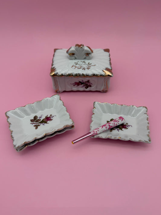 Footed rectangular fluted porcelain box with a gold crest handle and hand-painted floral sprays, beside two matching square scalloped ashtrays, one holding a glass cigarette pick with a floral handle, pink background.