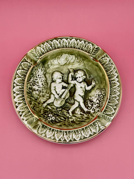 Round porcelain ashtray with raised cherub relief in olive and ivory tones with gold accents, pink background