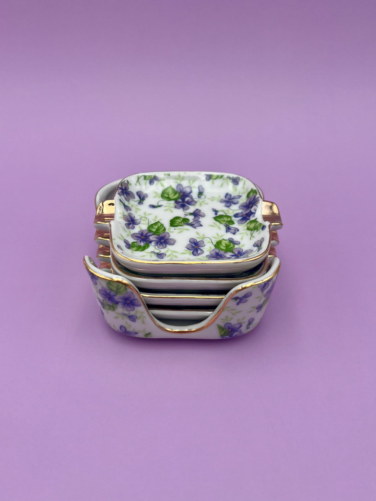 Stack of square porcelain ashtrays from the Felicity Set, each hand-painted with violet flowers and gold trim, purple background.