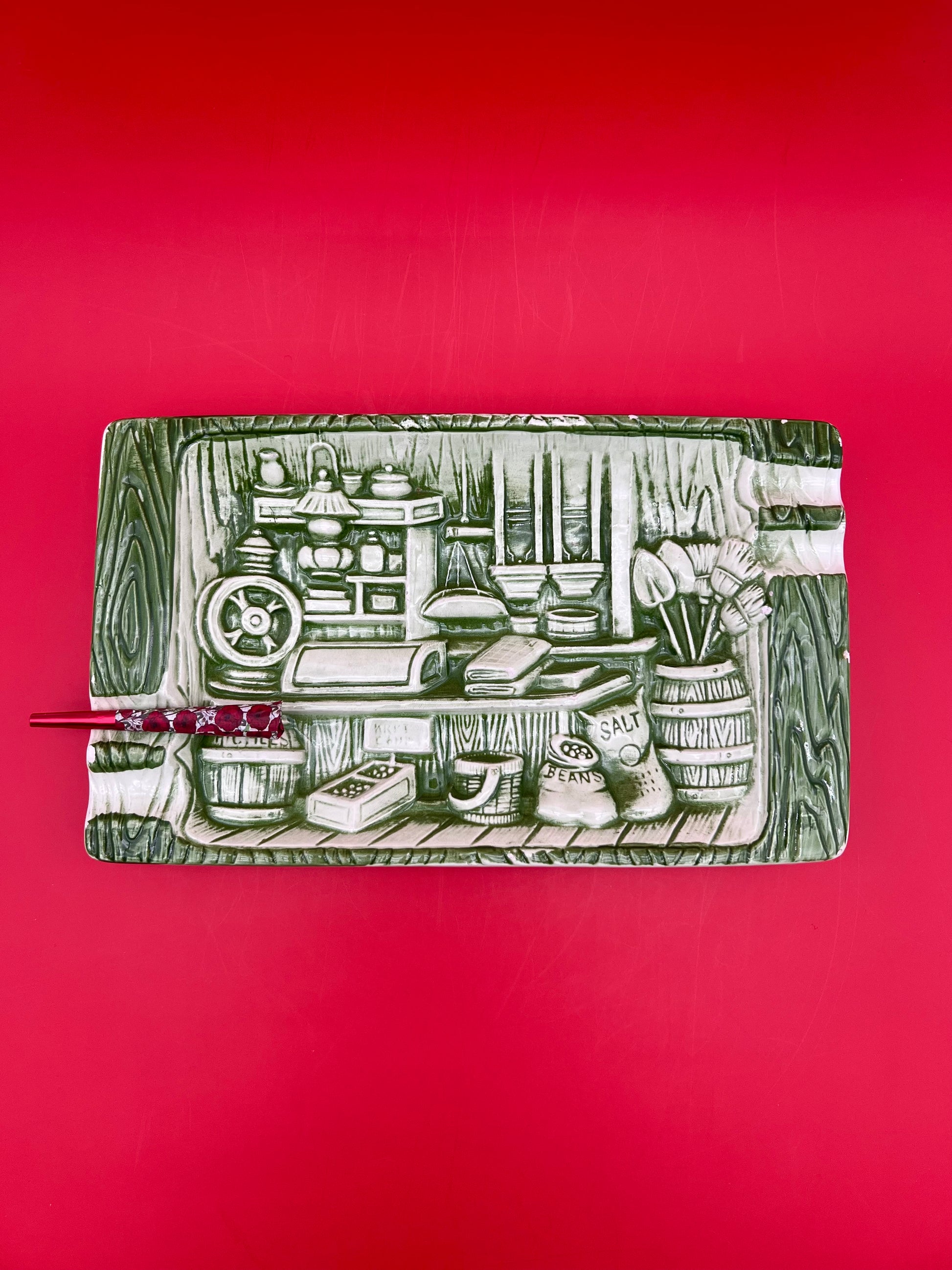 Rectangular sage green and white ceramic tray etched with a vintage kitchen pantry scene, glass cigarette pick resting across it, red background.