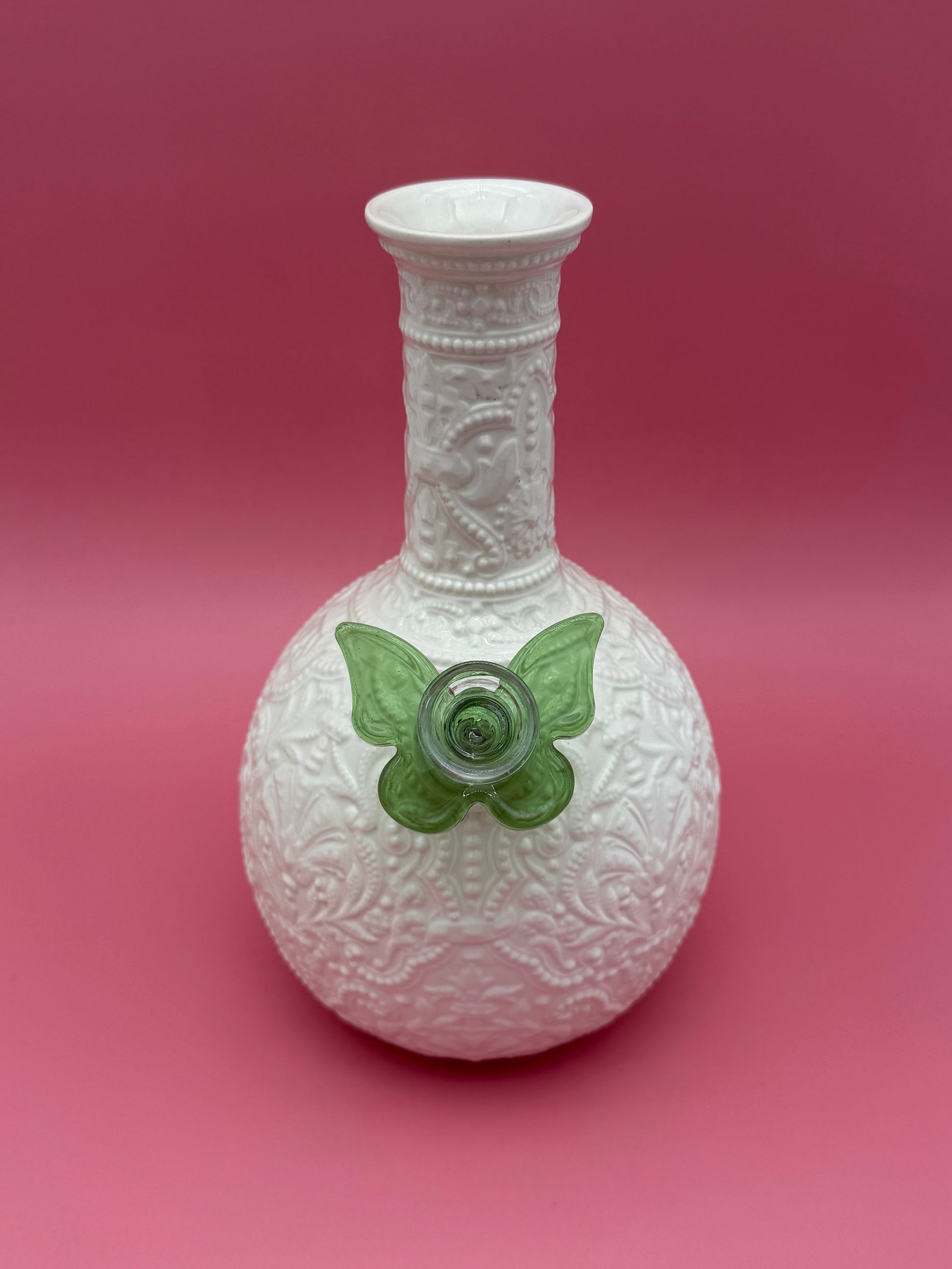 Front view of a white embossed porcelain bud vase water pipe with a heart-shaped bowl, green butterfly-shaped glass bowl piece attached at the side, magenta background.