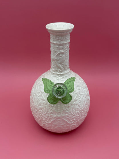 Front view of a white embossed porcelain bud vase water pipe with a heart-shaped bowl, green butterfly-shaped glass bowl piece attached at the side, magenta background.