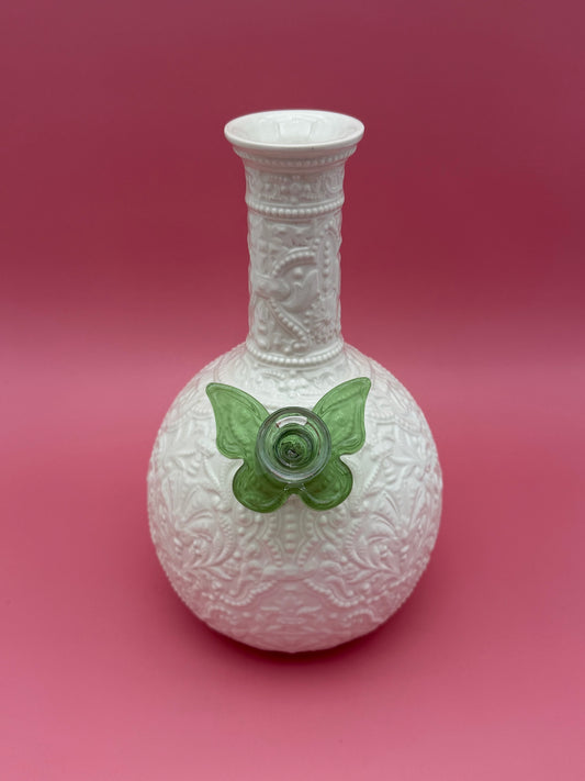 Front view of a white embossed porcelain bud vase water pipe with a heart-shaped bowl, green butterfly-shaped glass bowl piece attached at the side, magenta background.