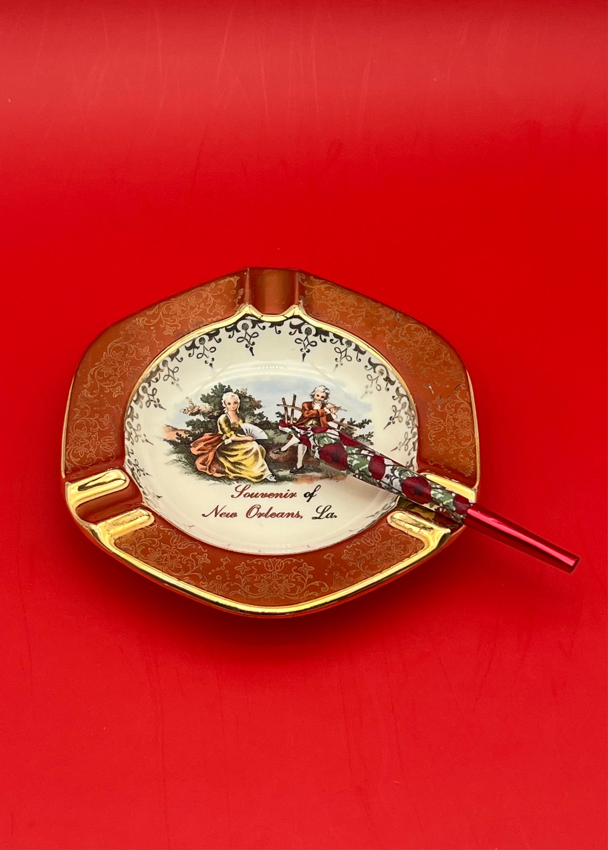 Same round scalloped ashtray with the painted Victorian courting couple scene and 'Souvenir of New Orleans, La.' text, now with a glass cigarette pick resting across it, red background.