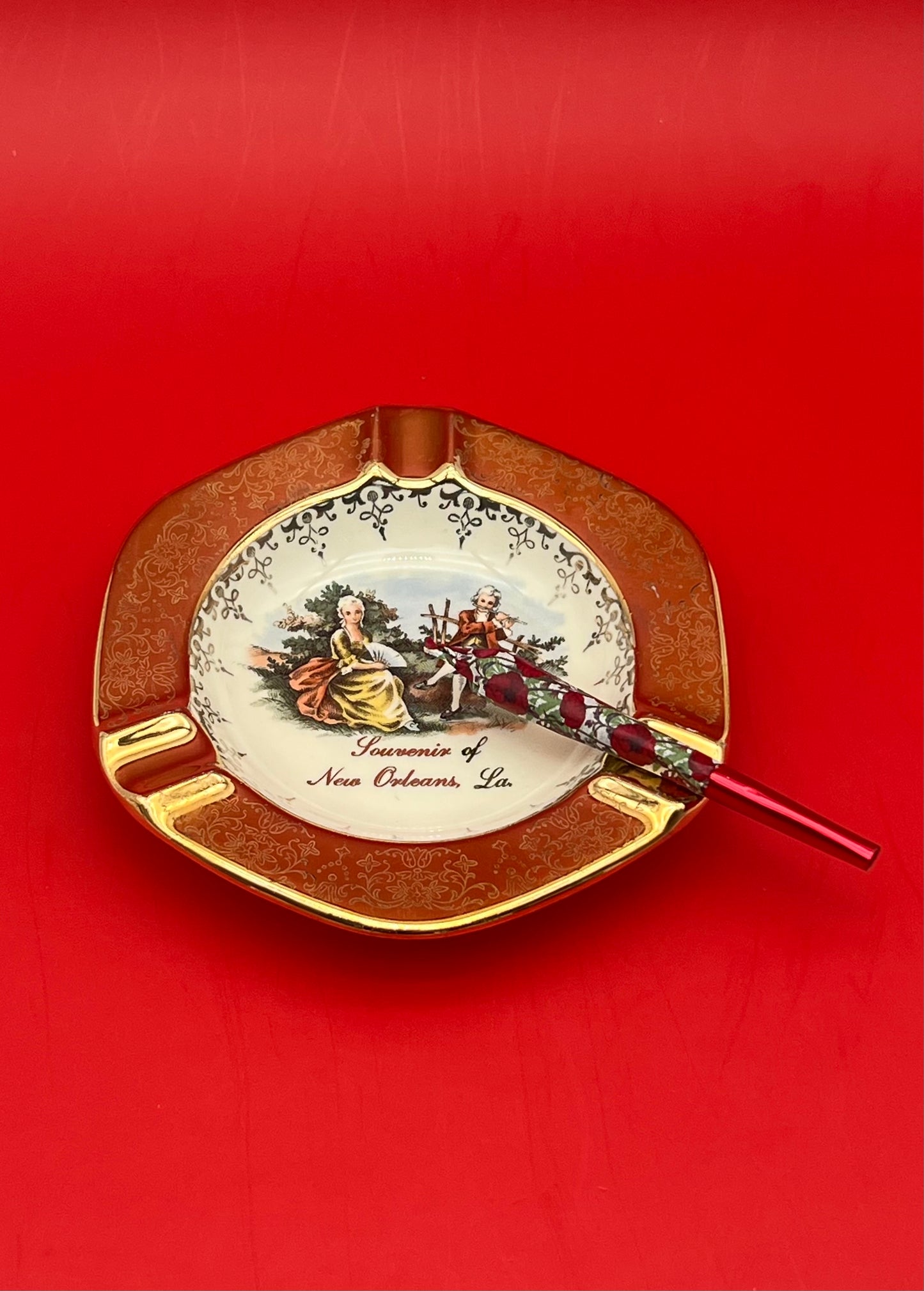 Same round scalloped ashtray with the painted Victorian courting couple scene and 'Souvenir of New Orleans, La.' text, now with a glass cigarette pick resting across it, red background.