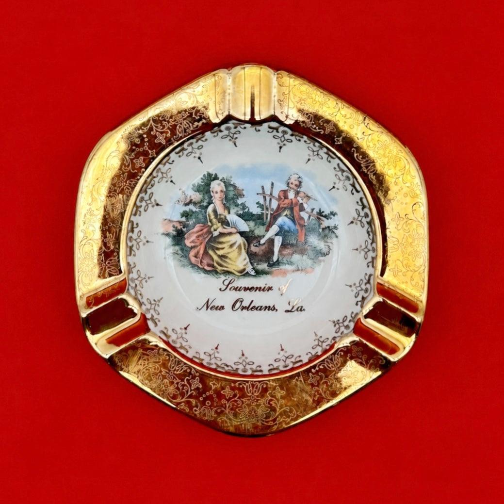 Round ceramic ashtray with an ornate gold scalloped rim and a hand-painted Victorian courting scene in the center, red background.