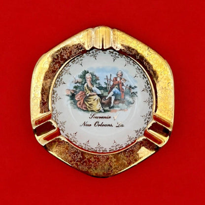 Round ceramic ashtray with an ornate gold scalloped rim and a hand-painted Victorian courting scene in the center, red background.