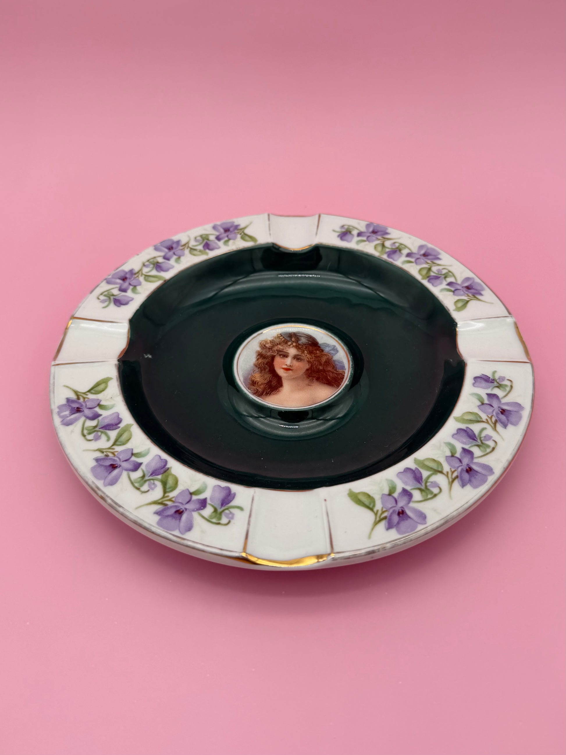 Round ashtray with a charcoal-gray bowl center and purple violet floral border, cameo portrait of a woman in the middle, pink background.