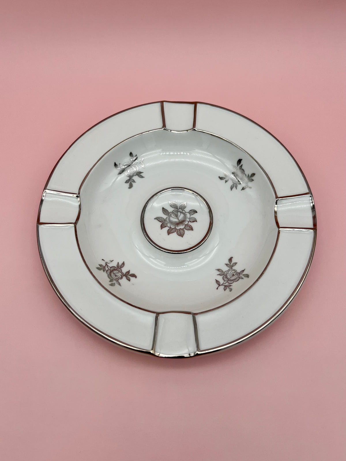 Round white and silver scalloped ashtray with small silver floral sprigs, no portrait, pink background.