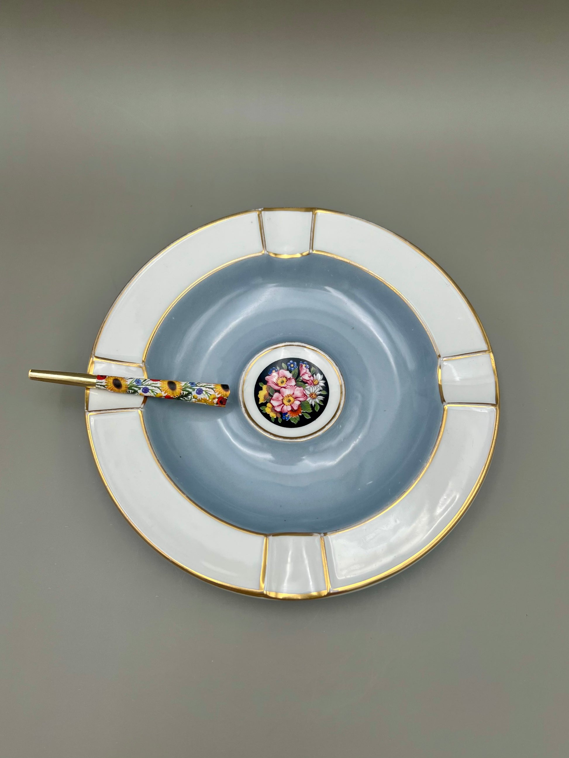 Same white and silver scalloped ashtray with a glass cigarette pick resting across it, blue-gray background.