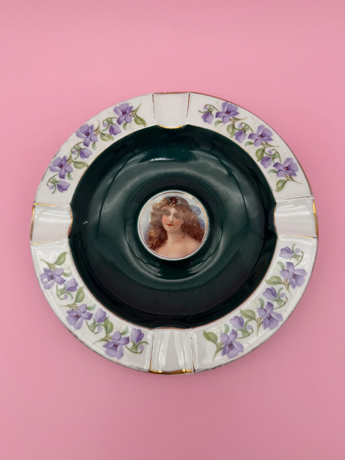 Round ceramic ashtray with a dark green rim, a cameo portrait of a woman in the center, and a purple floral border, pink background.