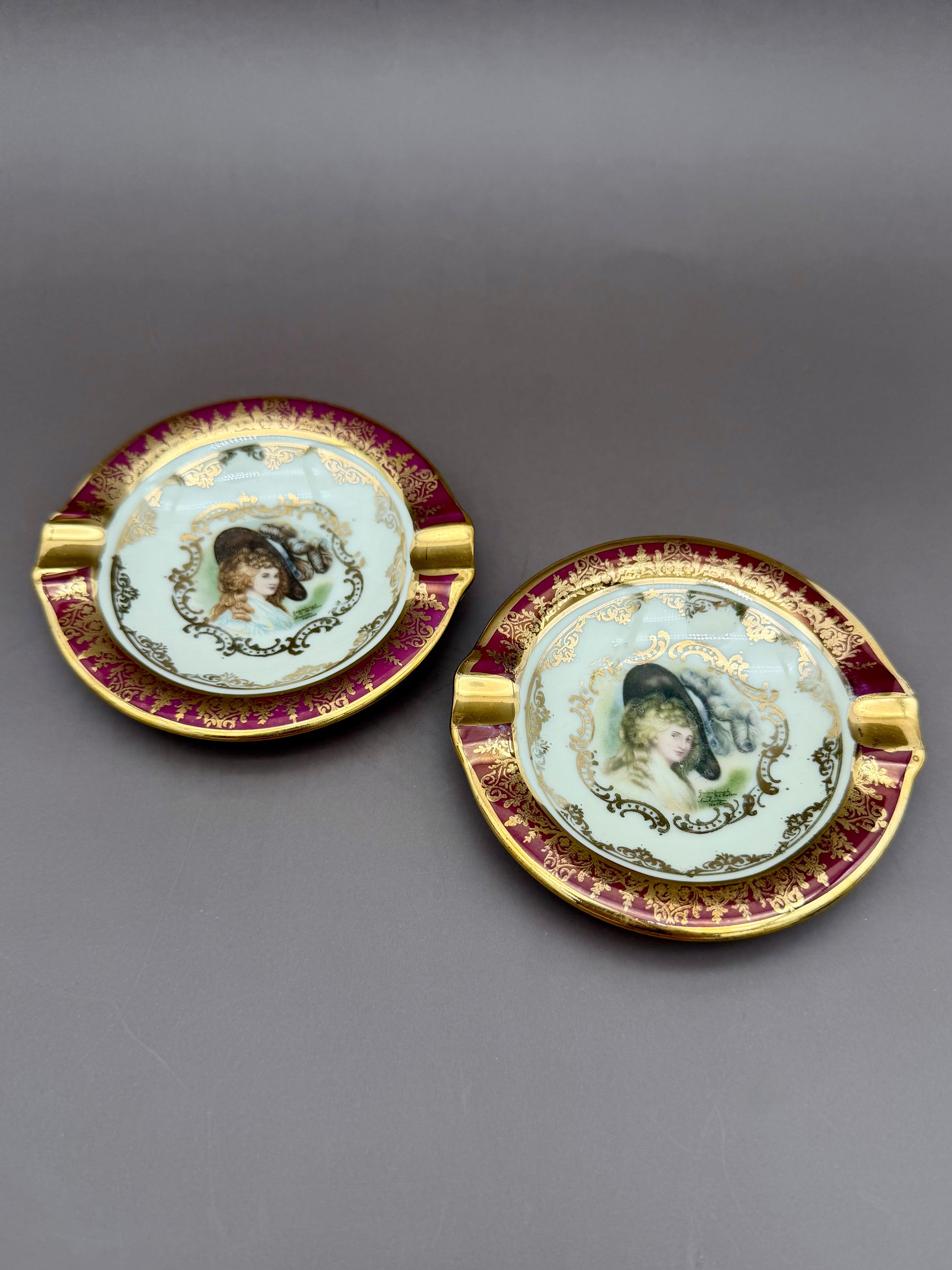 Two oval ashtrays from the Henrietta Set with burgundy and gold rims, each painted with a portrait medallion of a woman in a bonnet, overlapping arrangement, gray background.