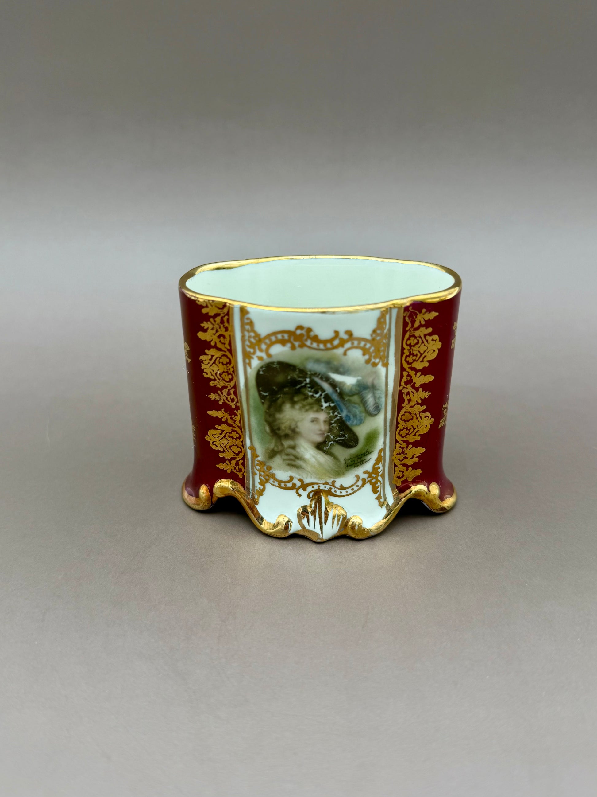 Front view of the Henrietta Set's cigarette holder cup showing a burgundy panel with a painted portrait medallion of a woman and gold scrollwork trim, gray background.