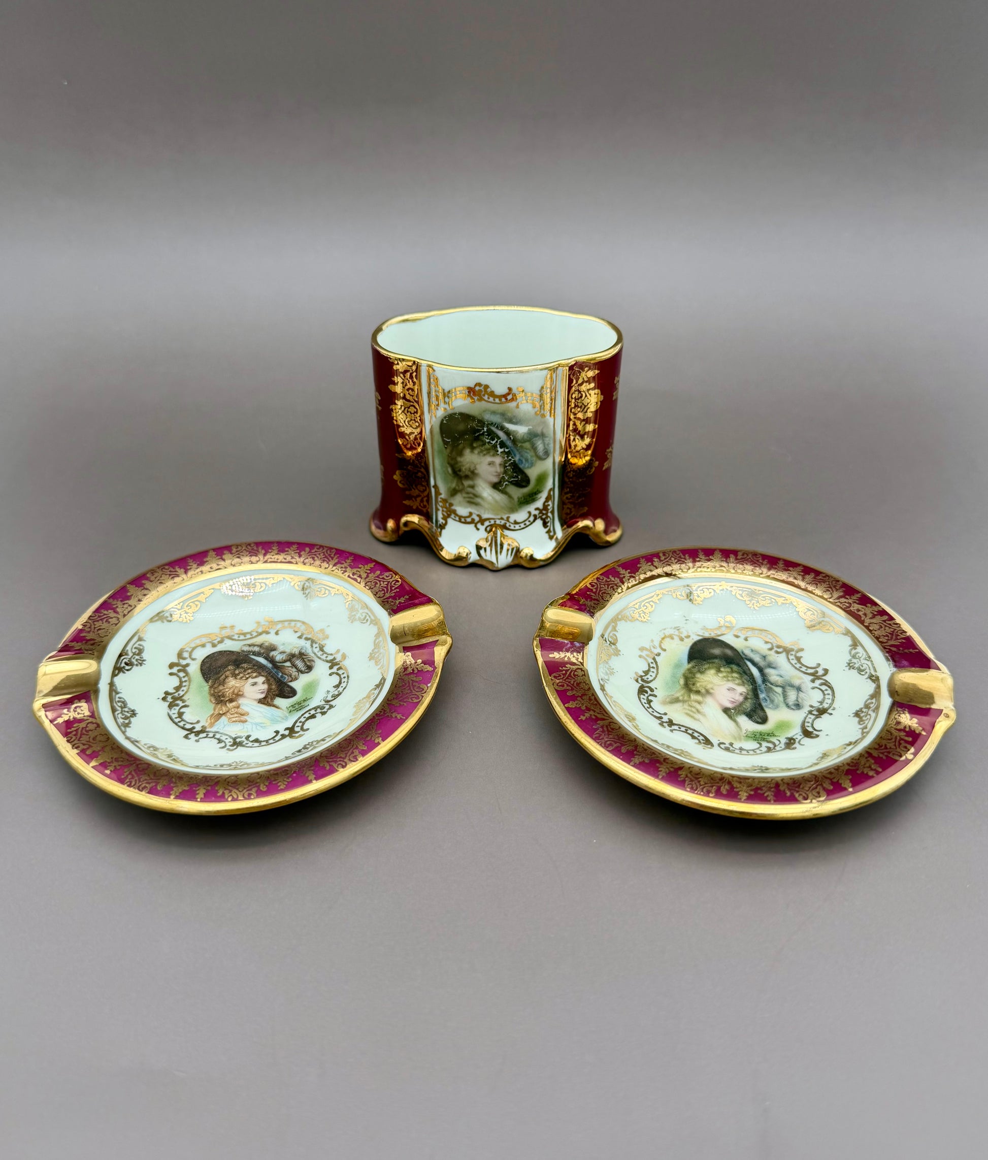 Henrietta Set featuring a cigarette holder cup and two oval ashtrays with burgundy and gold rims, each decorated with a painted portrait medallion, gray background.