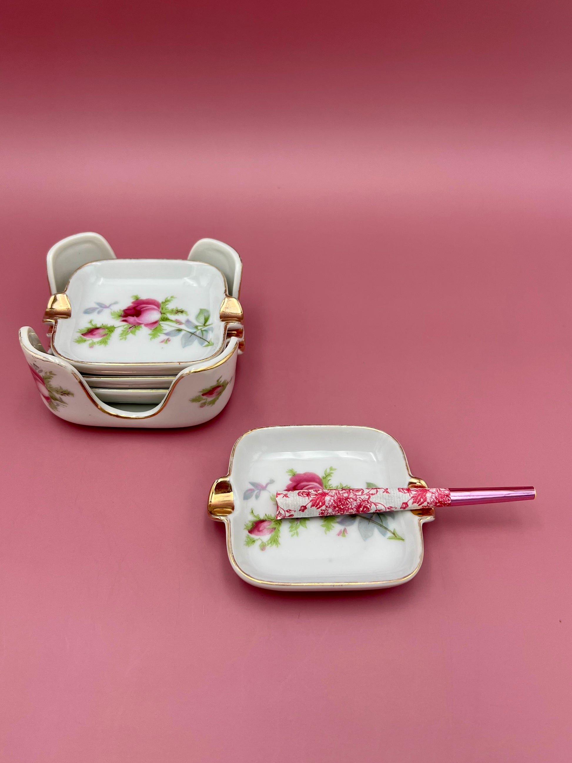 Two square white ashtrays with pink rose patterns and gold handles from the Isabella Set, one stacked pair in back and one with a glass cigarette pick resting across it in front, pink background.