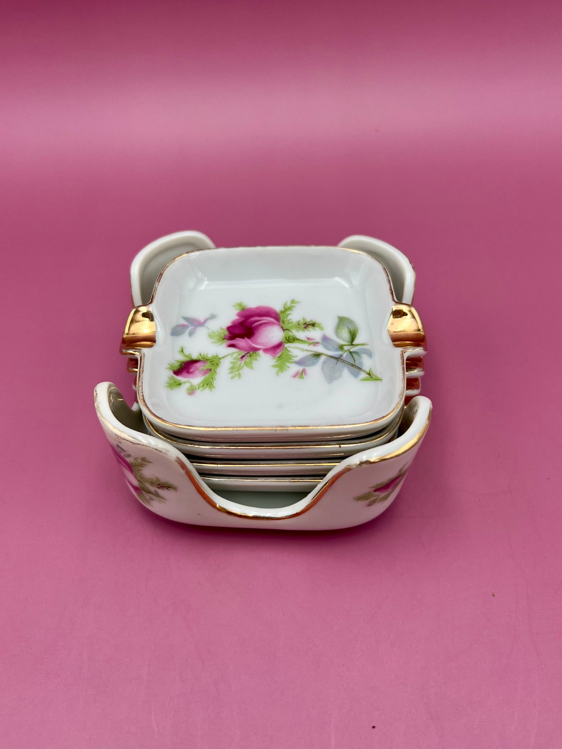 Stack of square white ashtrays with pink rose patterns and gold handles from the Isabella Set, pink background.