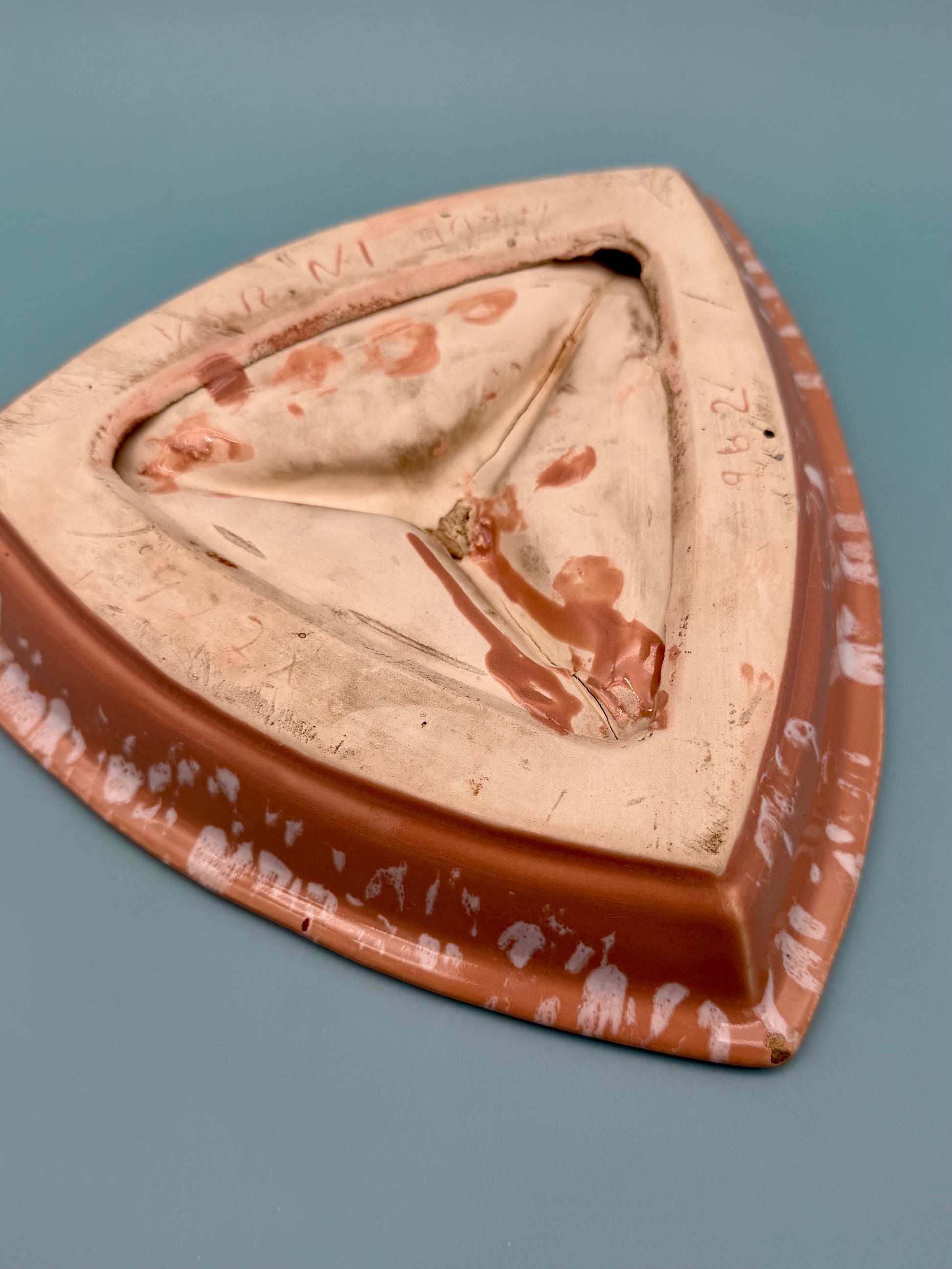 Same triangular terracotta ashtray with speckled glaze, viewed from a different angle showing its three cigarette rests, gray background.