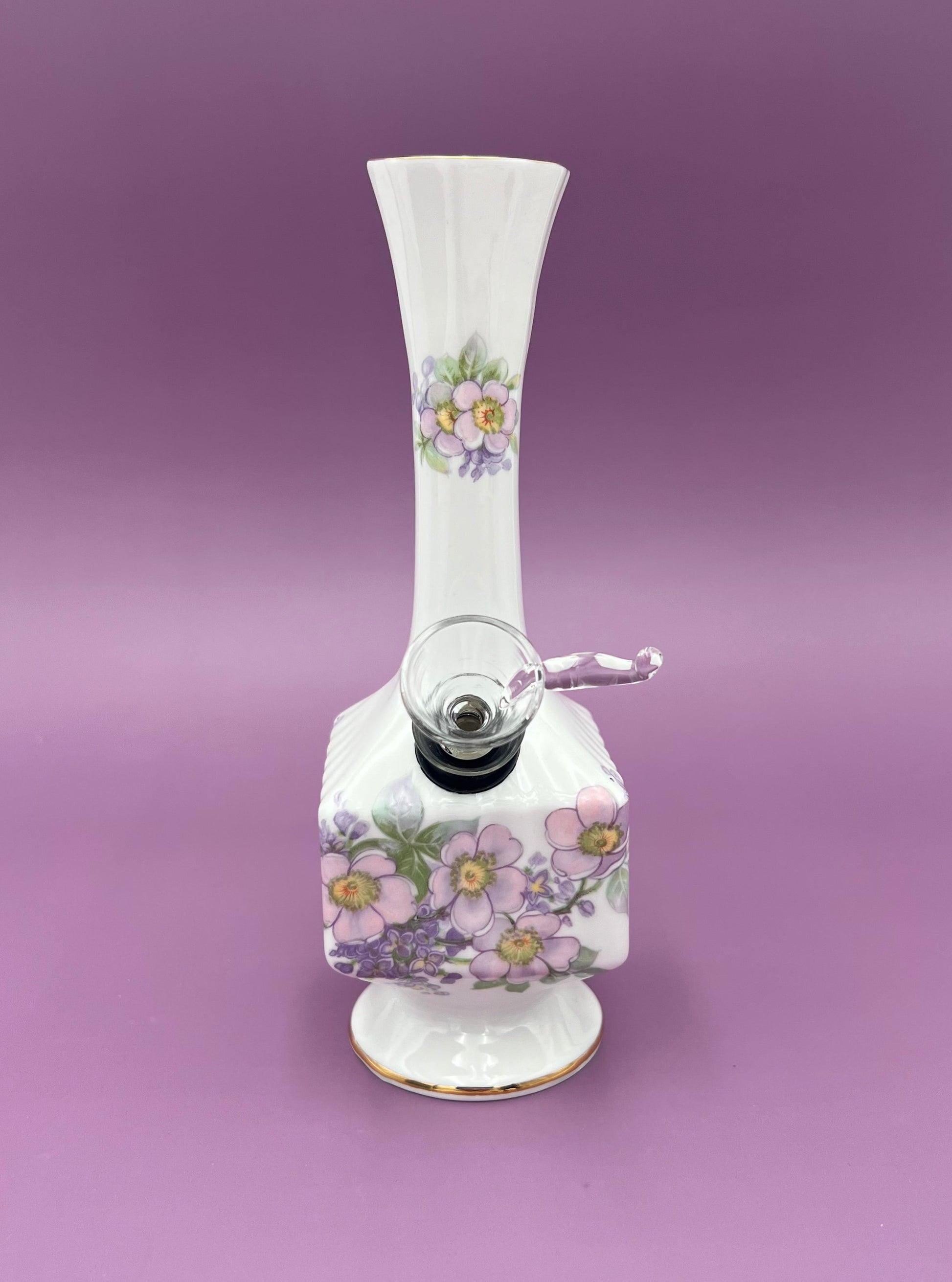 Front view of a white porcelain bud vase water pipe with a wreath of purple flowers and green leaves, gold trim, glass fittings on the side, purple background.