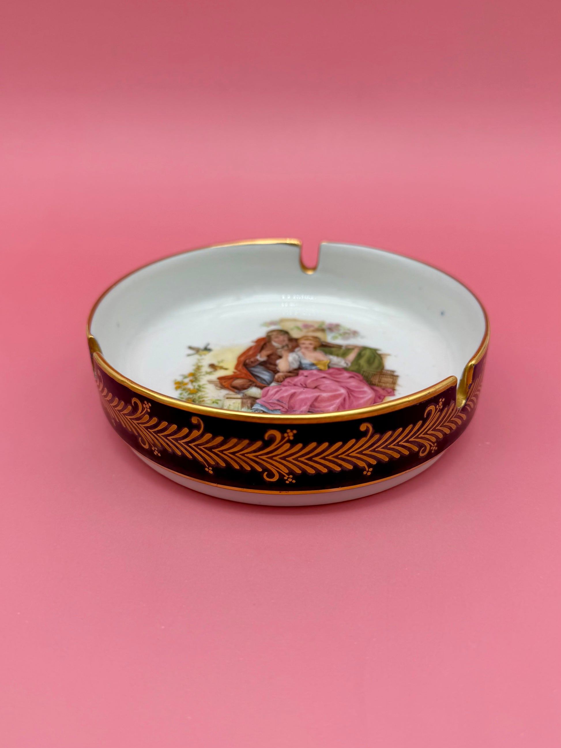 Interior of the same round ashtray showing the painted Victorian courting couple scene framed by a dark rim with gold fern-leaf border, pink background.