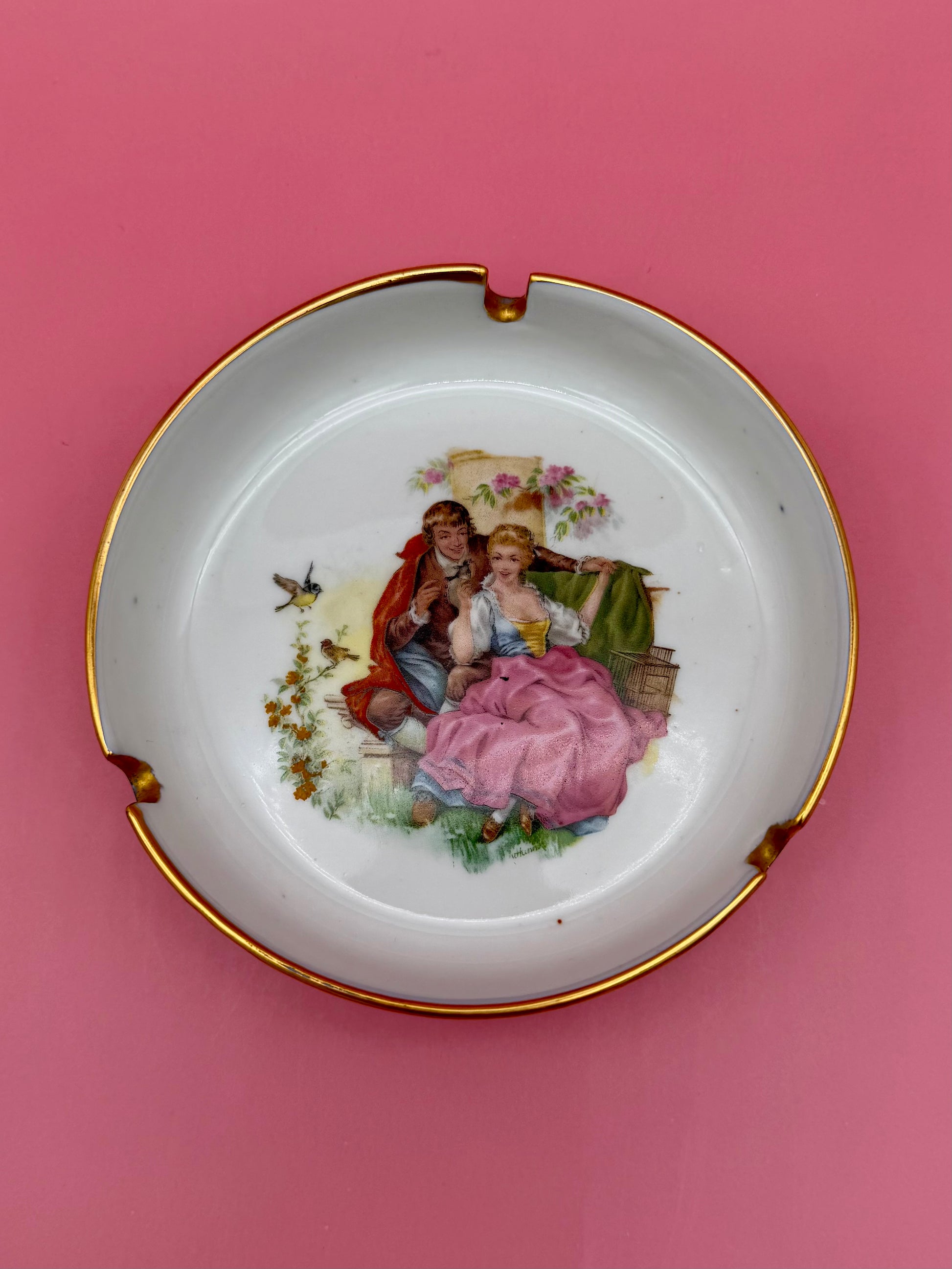 Round white ceramic ashtray with a gold rim and a hand-painted Victorian courting couple scene in the center, pink background.