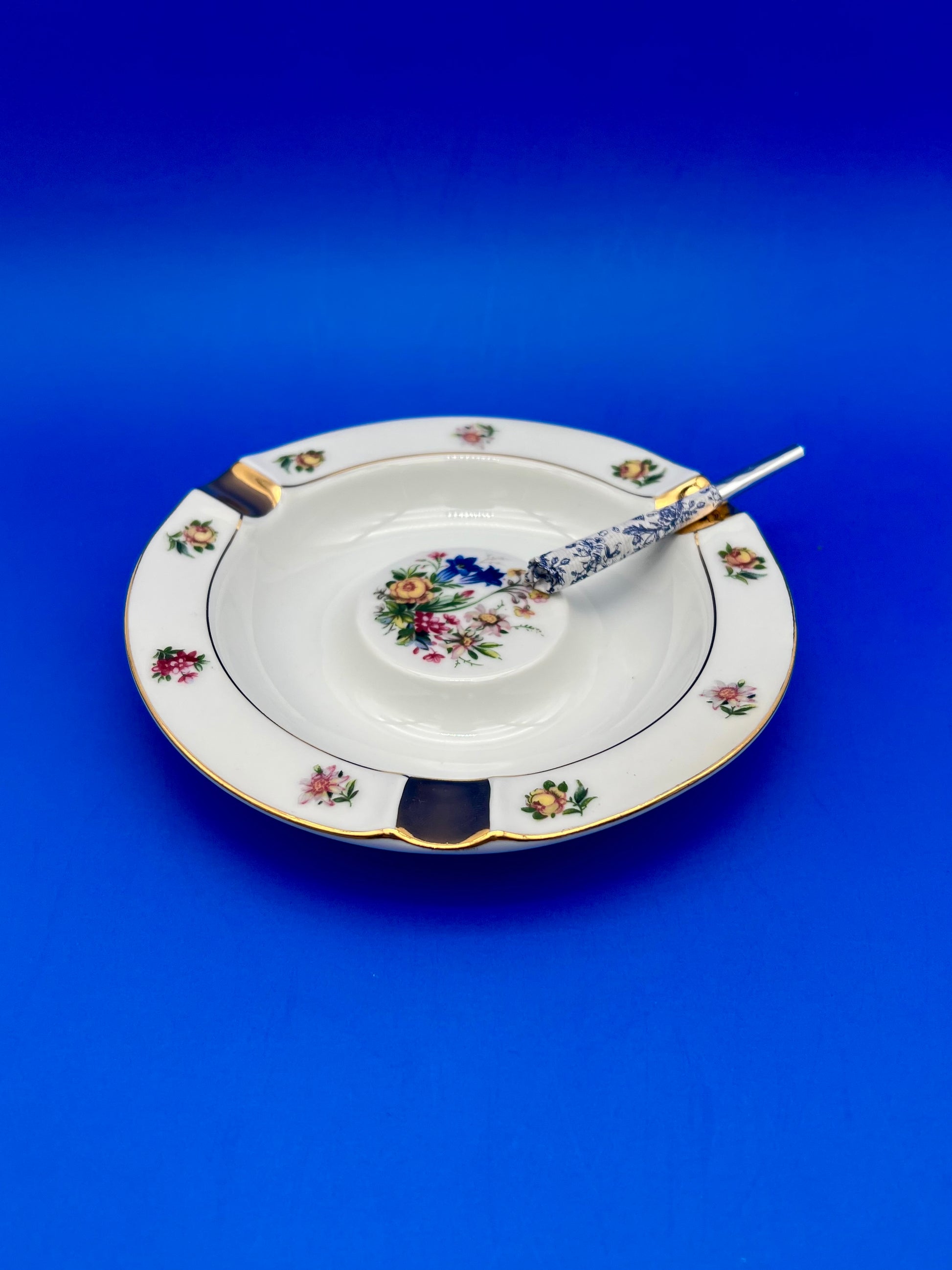 Same round gold-rimmed floral ashtray with a glass cigarette pick resting across it, blue background.