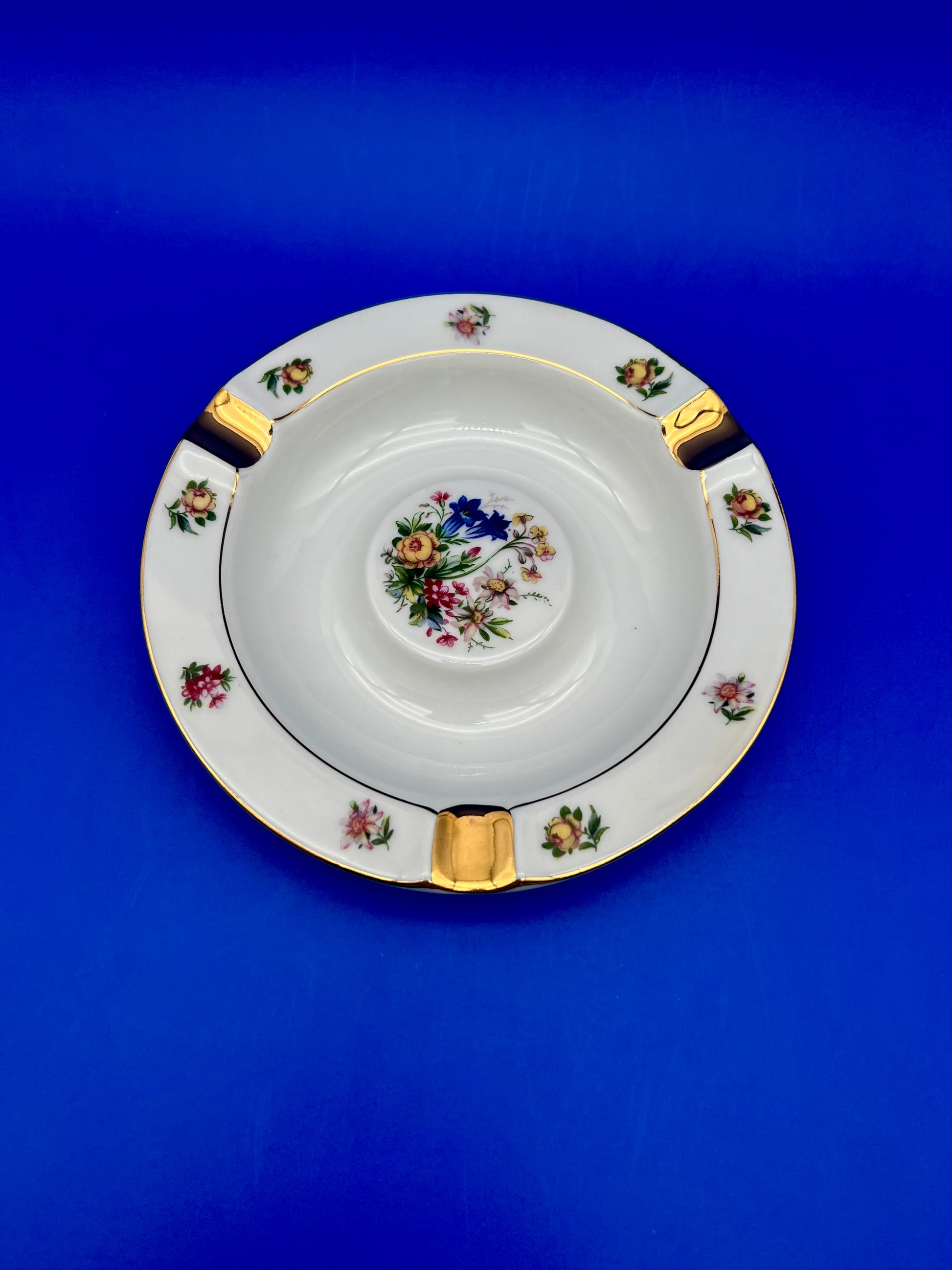 Round white ceramic ashtray with a gold rim and a colorful hand-painted floral bouquet in the center, blue background.