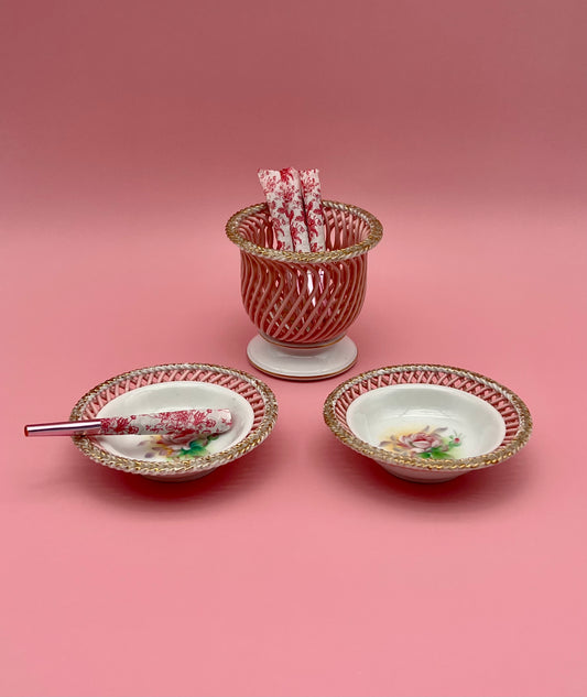 Lauren Set featuring a cylindrical holder cup and two round bowls with a gold lattice rim and pink floral pattern, glass cigarette picks resting in them, pink background.