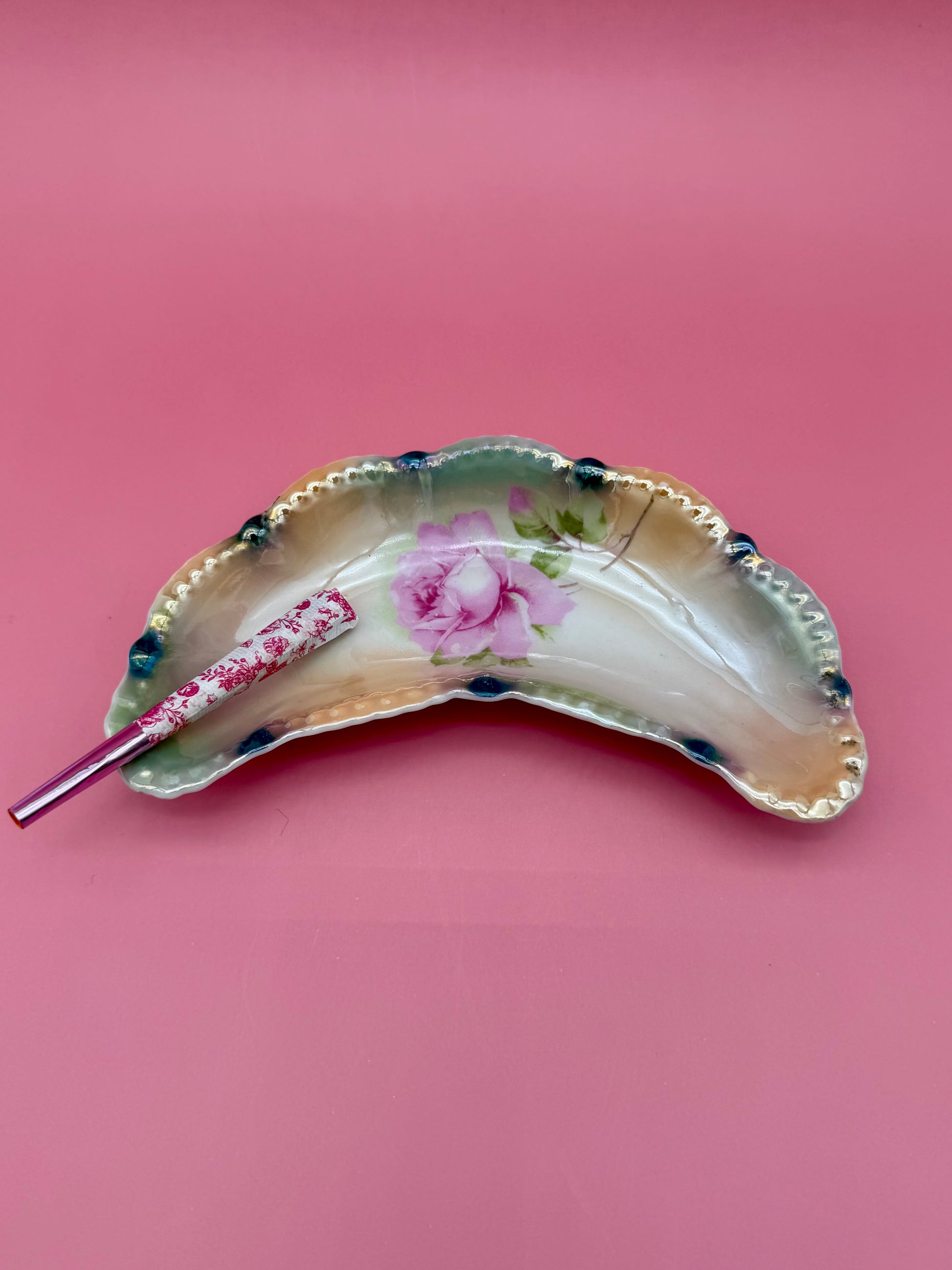 Crescent-shaped iridescent lustreware ashtray with a beaded edge and hand-painted pink rose design, glass cigarette pick resting across it, pink background.