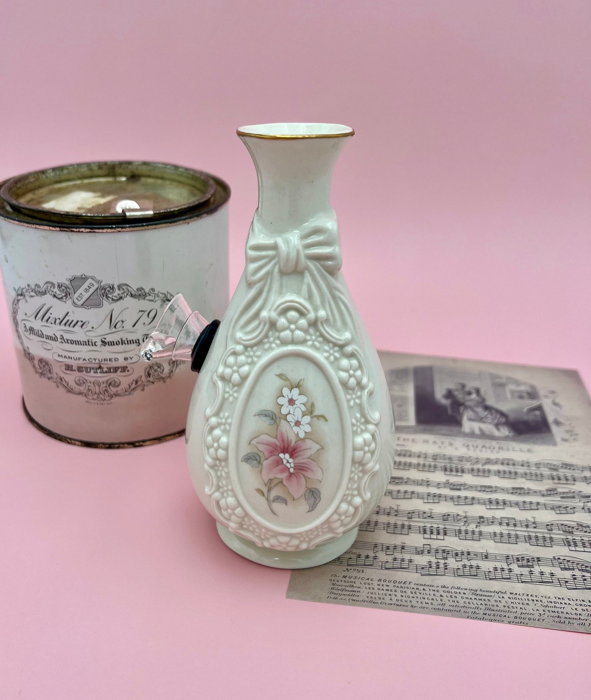 Louise water pipe styled next to a vintage tin and antique sheet music, showing the embossed bow and pink floral medallion in a decorative vignette.