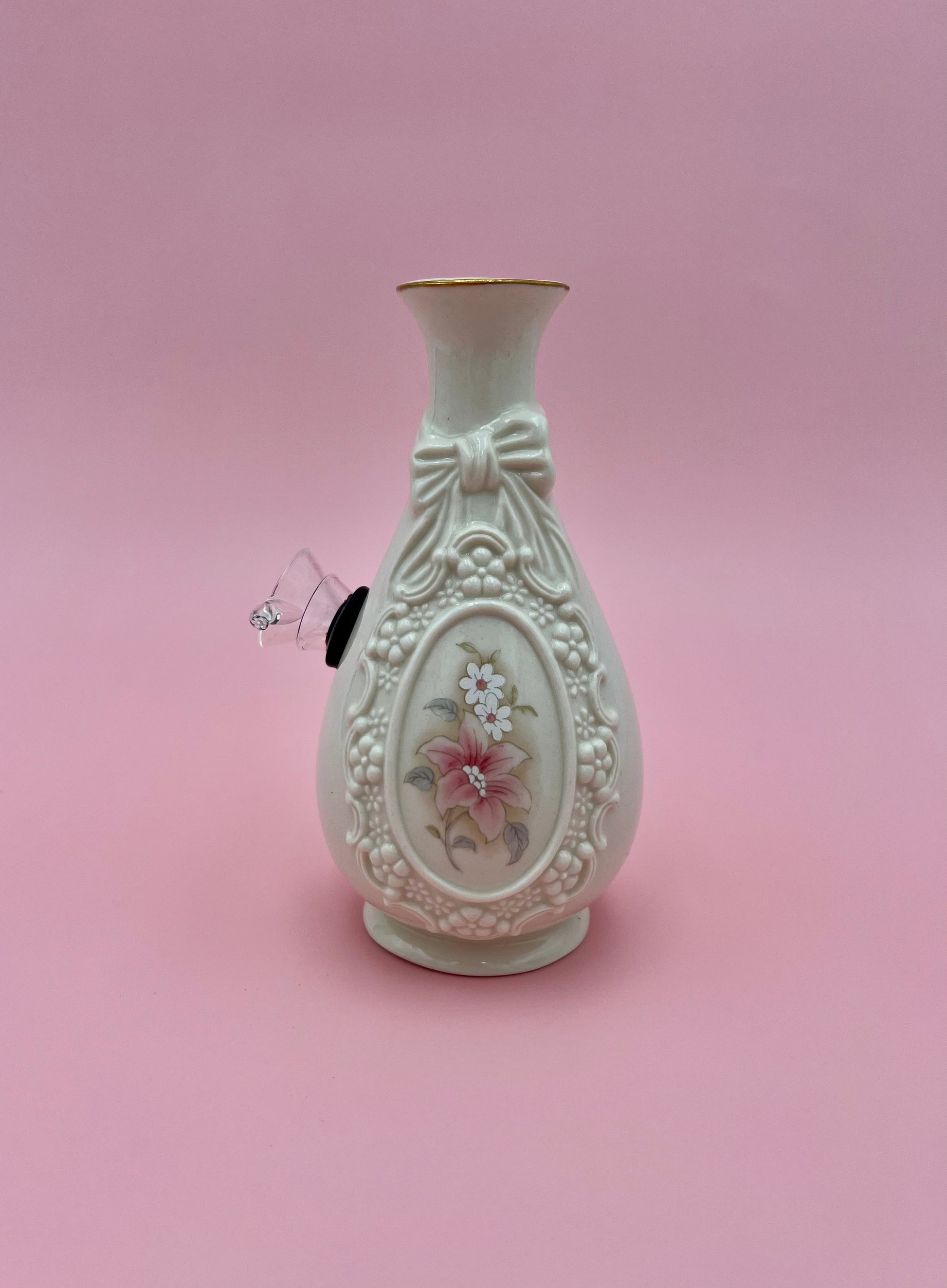 Front view of a cream porcelain bud vase water pipe with an embossed bow and ribbon design framing a pink rose medallion and gold rim, clear glass bowl piece fitted on the side, pink background.