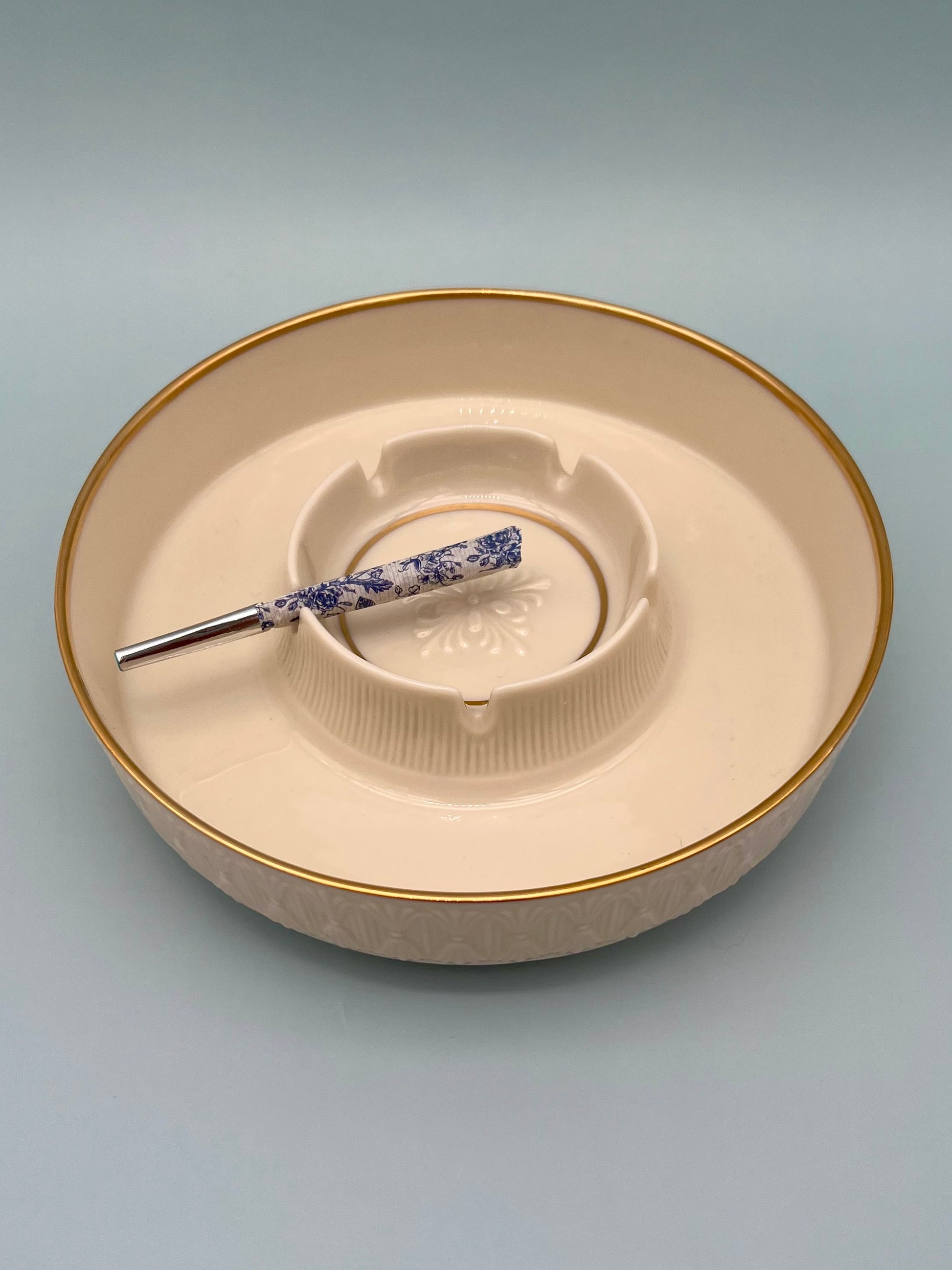 Round peach ceramic ashtray with a ribbed rim, glass cigarette pick with a floral patterned handle resting across it, blue-gray background.