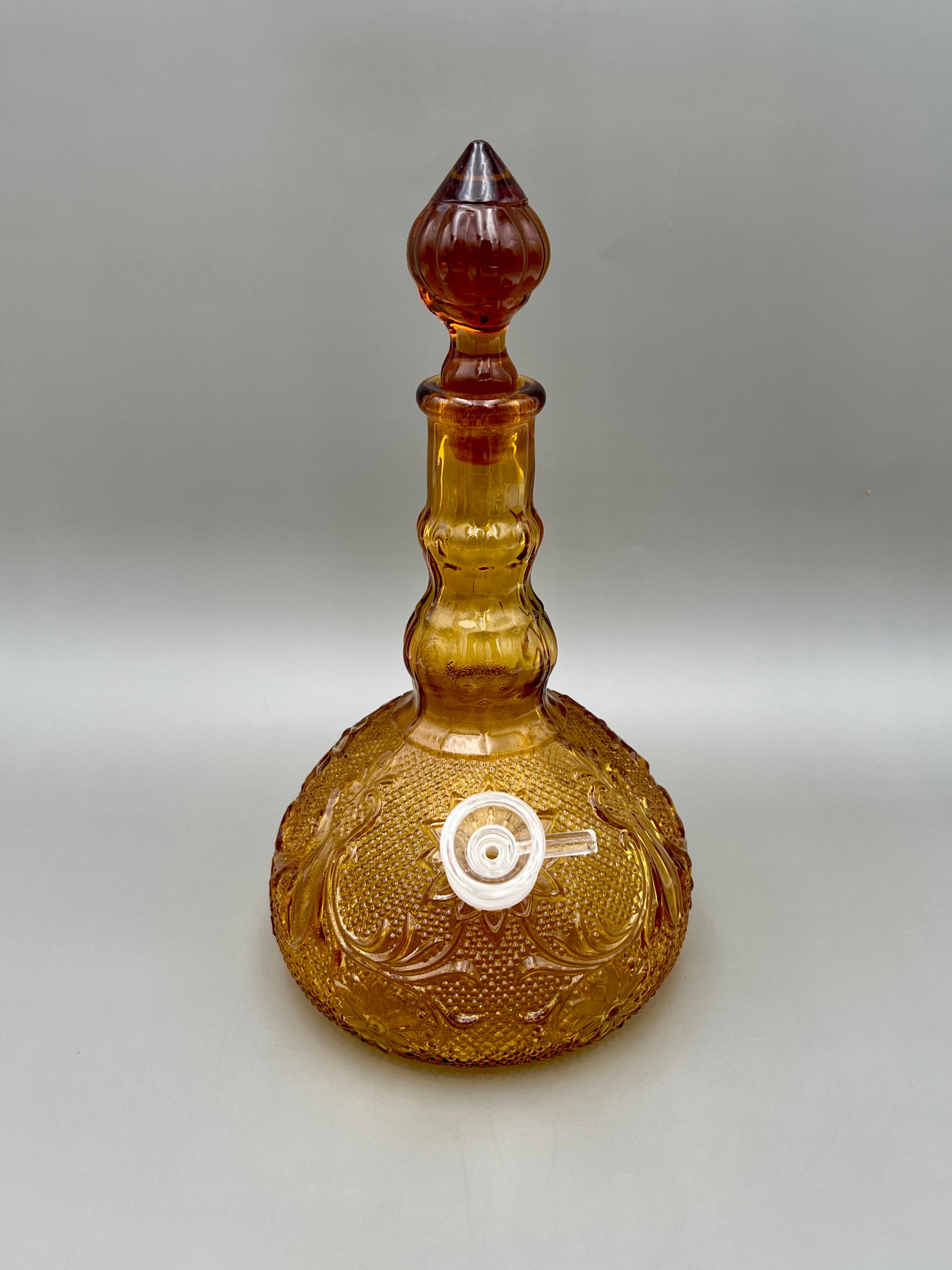 Amber pressed-glass genie-bottle decanter with a diamond-point pattern and faceted stopper in place, glass mouthpiece attached near the neck, gray background