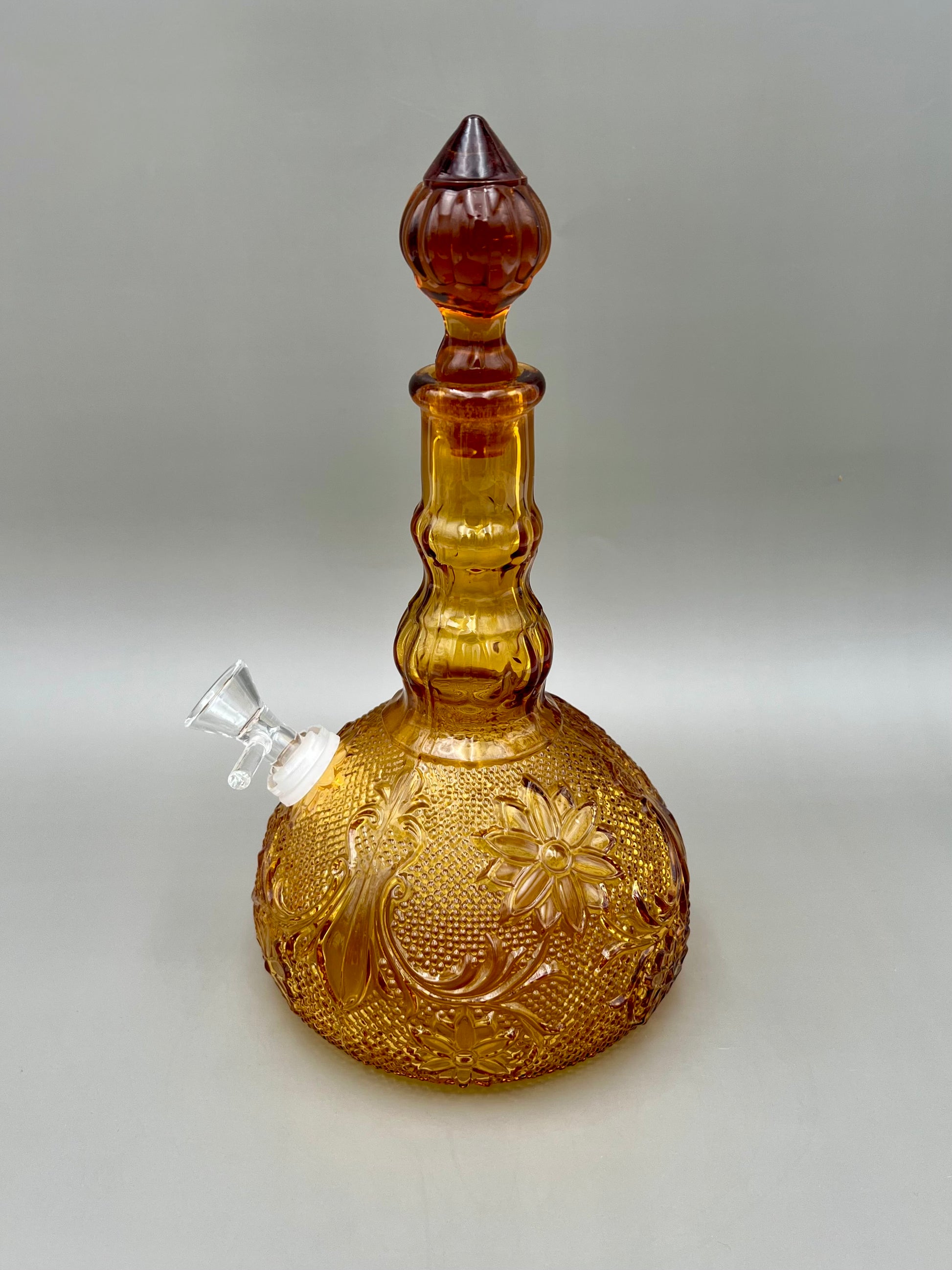 Same amber pressed-glass decanter with a white ceramic mouthpiece attachment at the neck and stopper in place, gray background