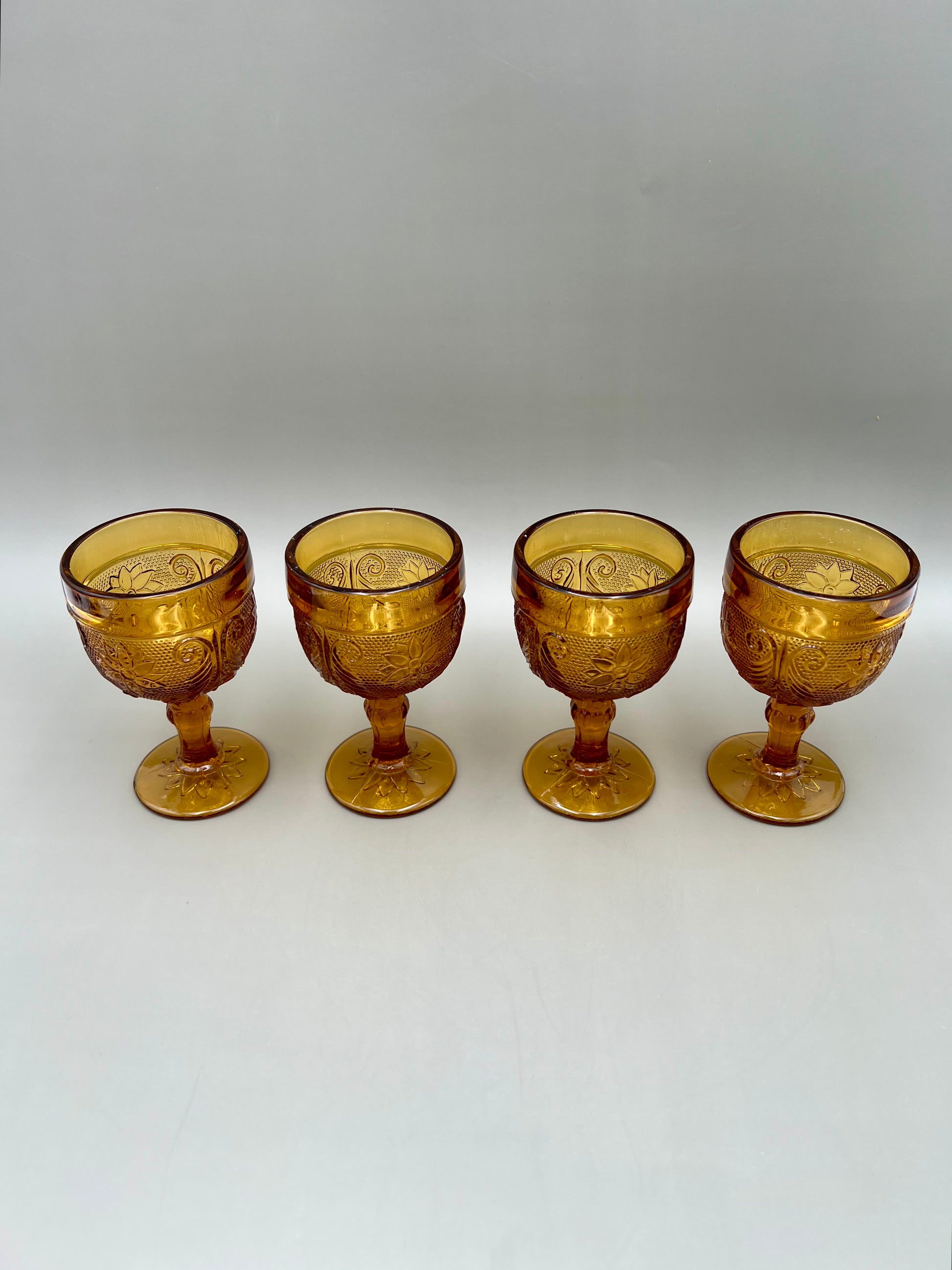 Four matching amber pressed-glass cordial goblets lined up in a row, gray background
