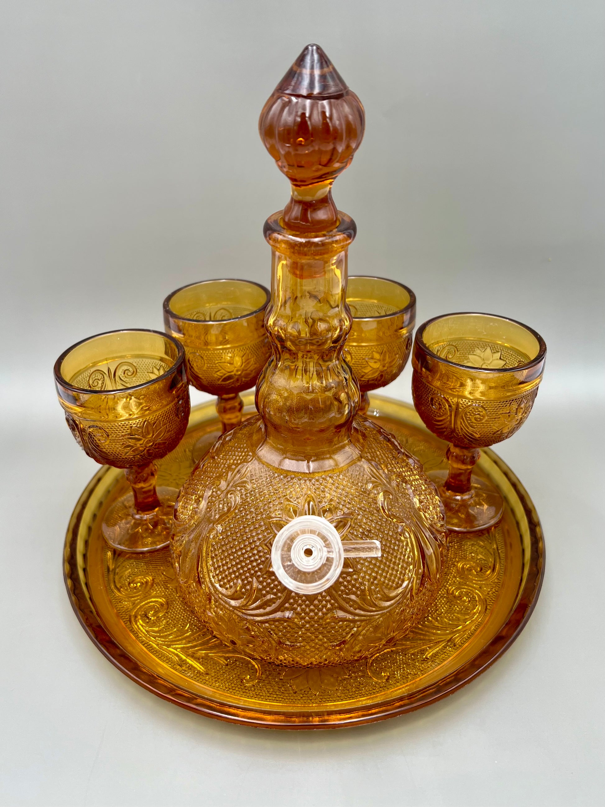 Mabel Set water pipe display featuring an amber pressed-glass decanter with stopper, a matching serving tray, and four small cordial glasses, against a light gray background.