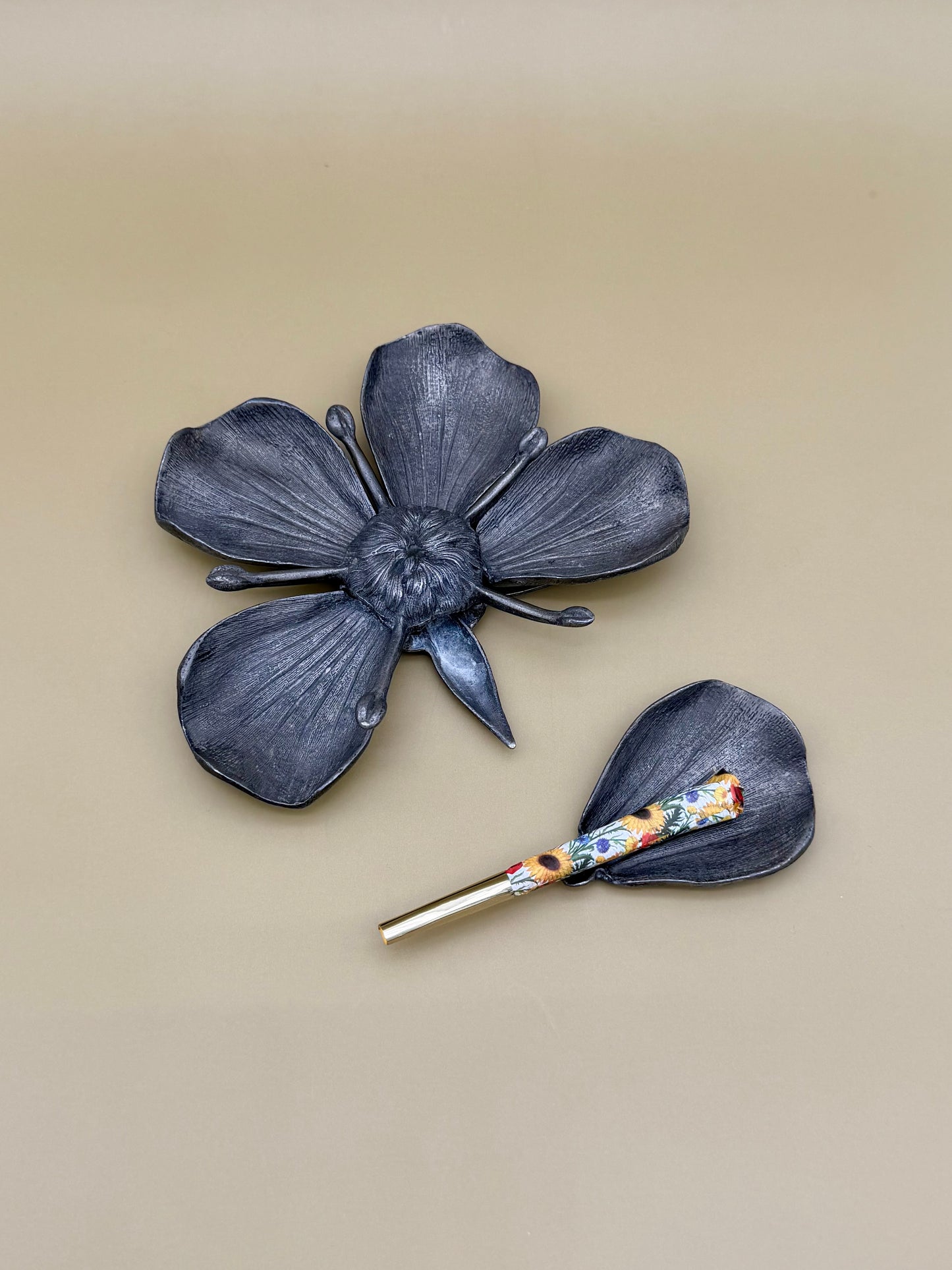 Silver marigold flower-shaped dish with five petals, paired with a small teardrop-shaped petal dish holding a floral-patterned glass cigarette pick, tan background.