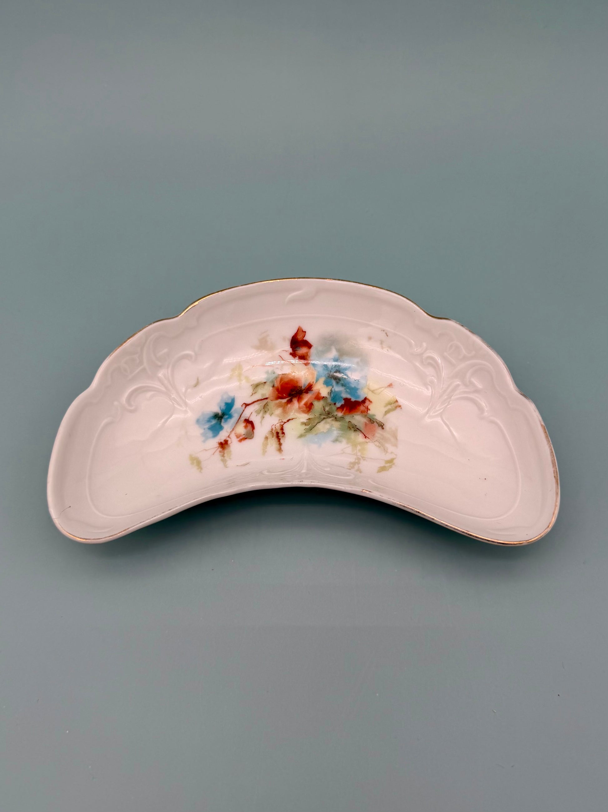 Crescent-shaped blush pink ceramic ashtray with an embossed rim and a hand-painted floral design, gray background.